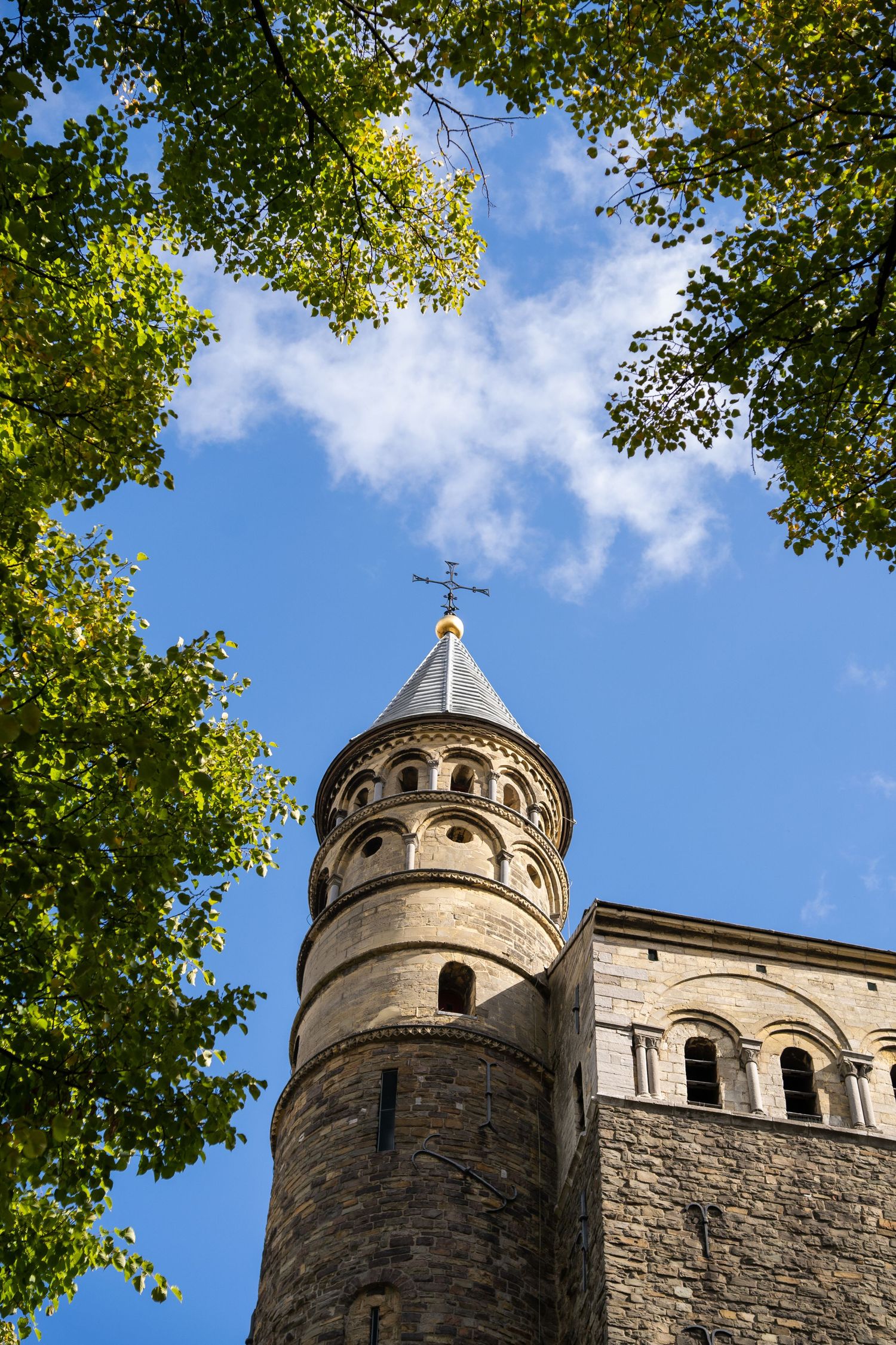 Historic Romanesque church tower framed by green trees against a blue sky with white clouds.