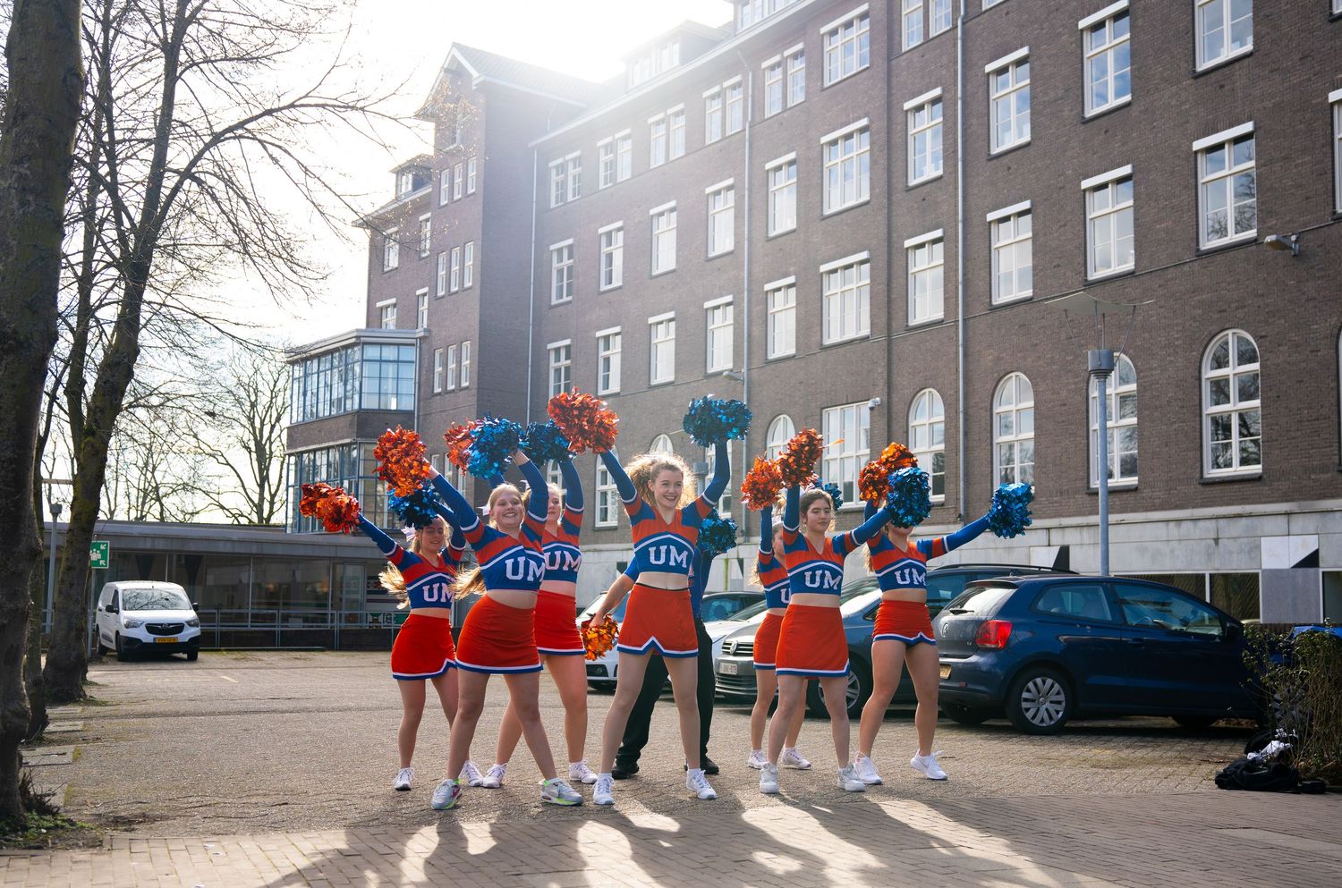 Cheerleaders in red and blue uniforms perform outside a brick university building on a sunny day.