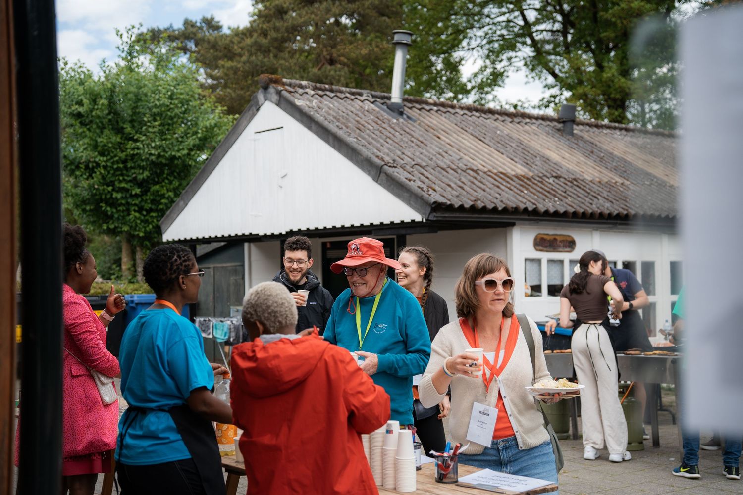 Volunteers and attendees socialize and share refreshments outside a rustic building at an outdoor event.