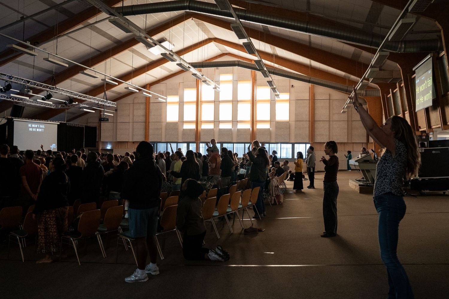 Congregation raising hands in worship inside a large wooden hall with high ceilings and natural light.