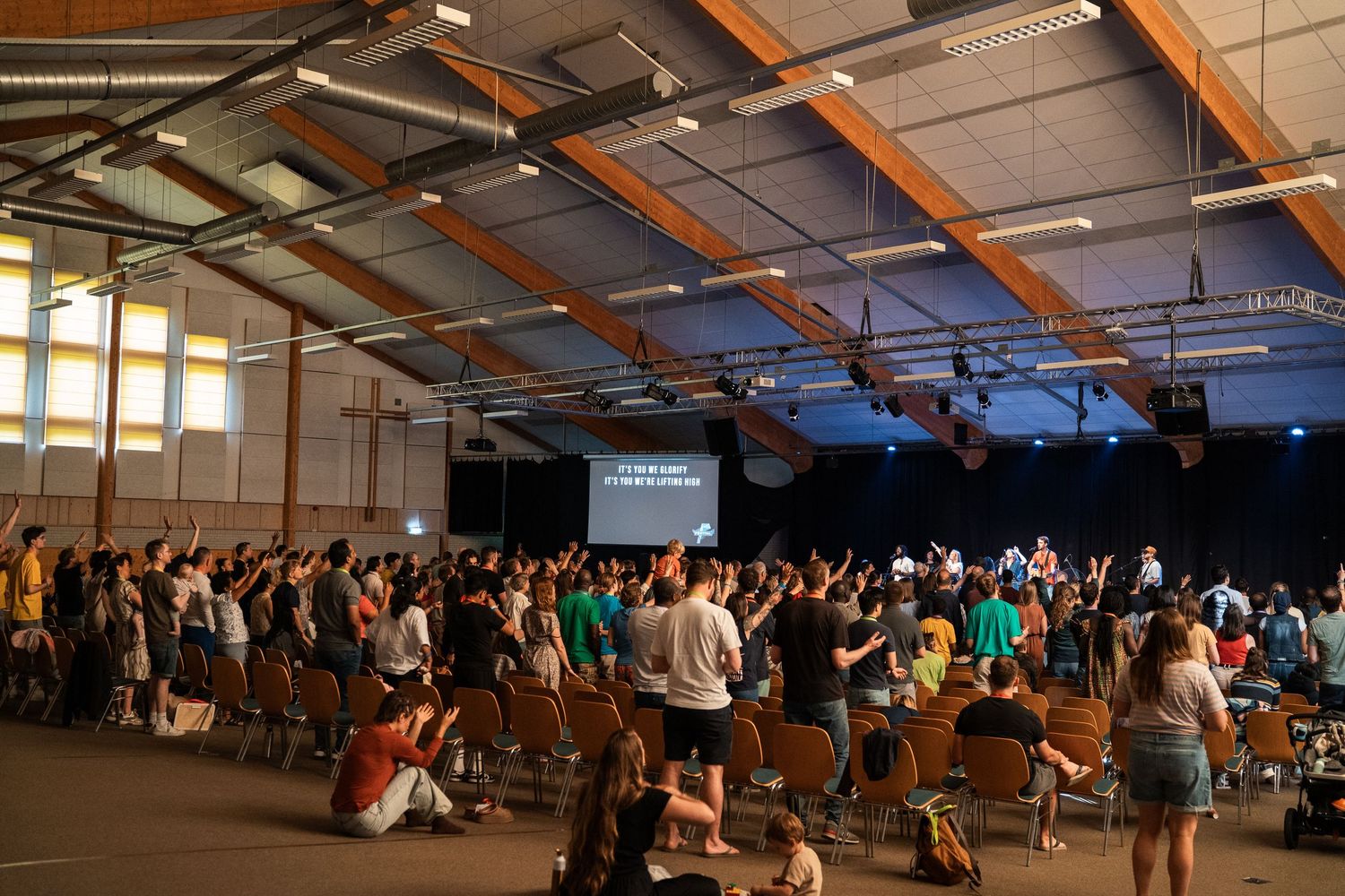 Large congregation raises hands in worship during a Christian youth conference in a spacious indoor hall.