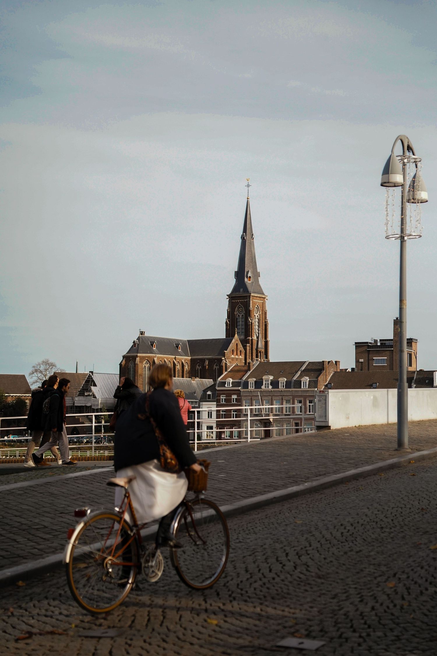 Cyclist riding over a cobblestone bridge toward a Gothic church steeple in a European city.