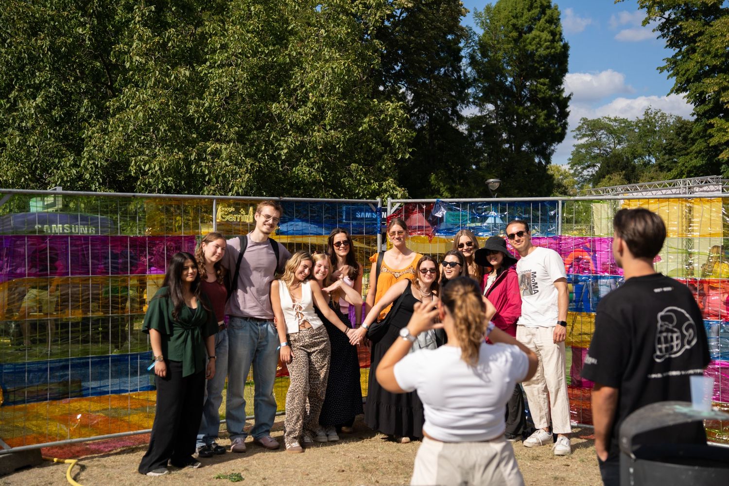 Group of smiling people posing for a photo in front of a colorful mosaic wall at an outdoor festival.