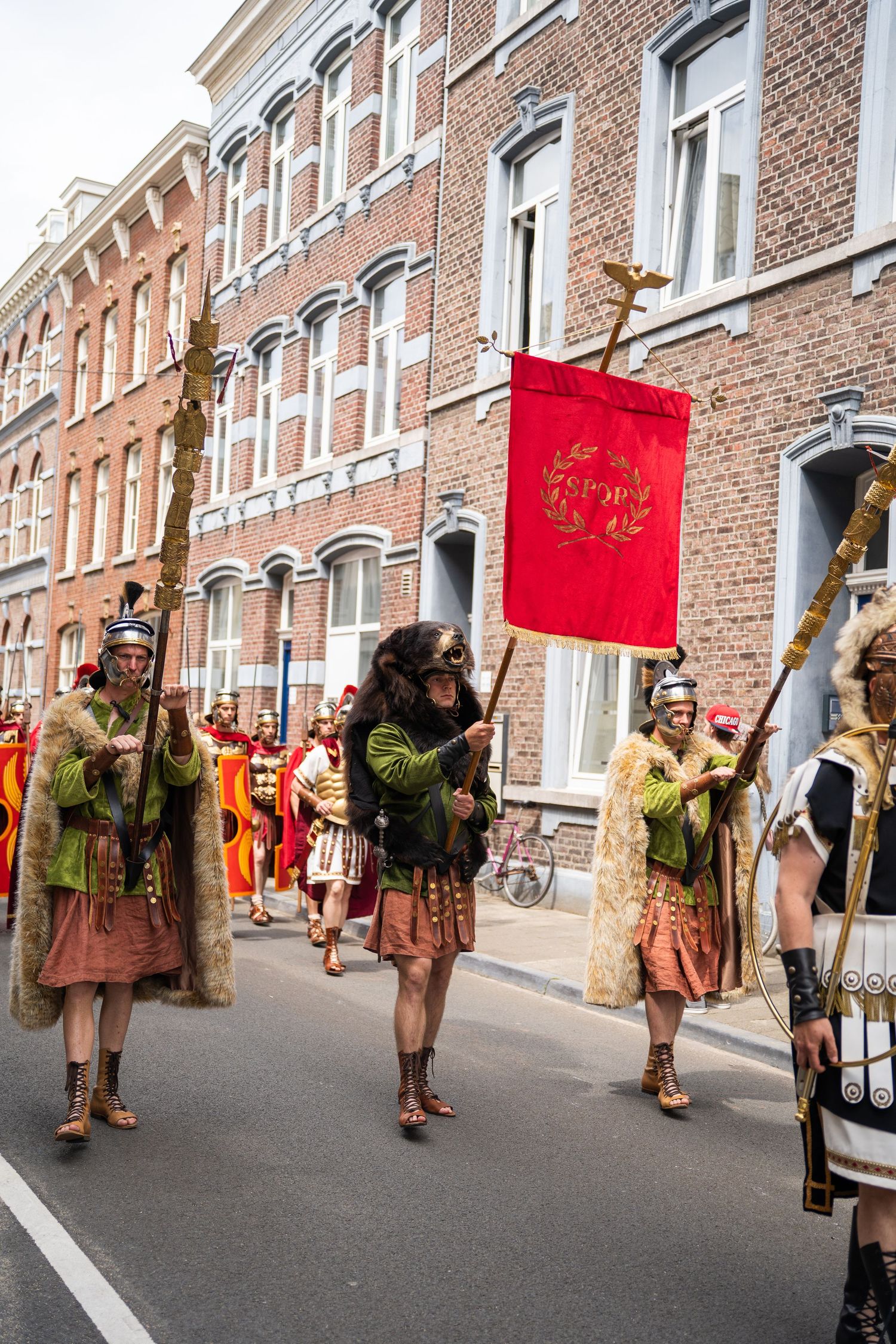 Performers in Roman soldier costumes carry a red banner in a historical street procession.