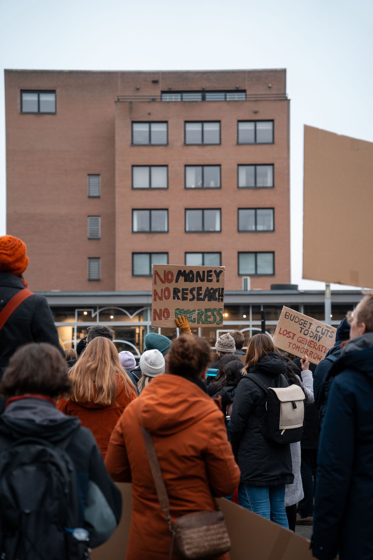 Crowd of protesters at a rally holding a sign reading No Money No Research No Progress against budget cuts.