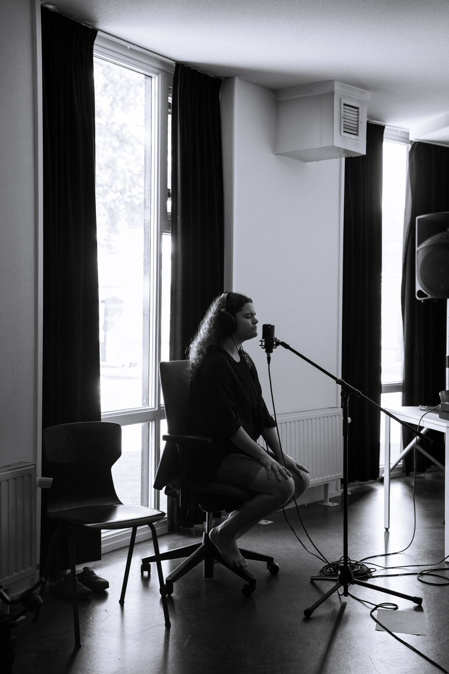 Black and white image of a woman with long hair singing into a microphone while seated in a studio.