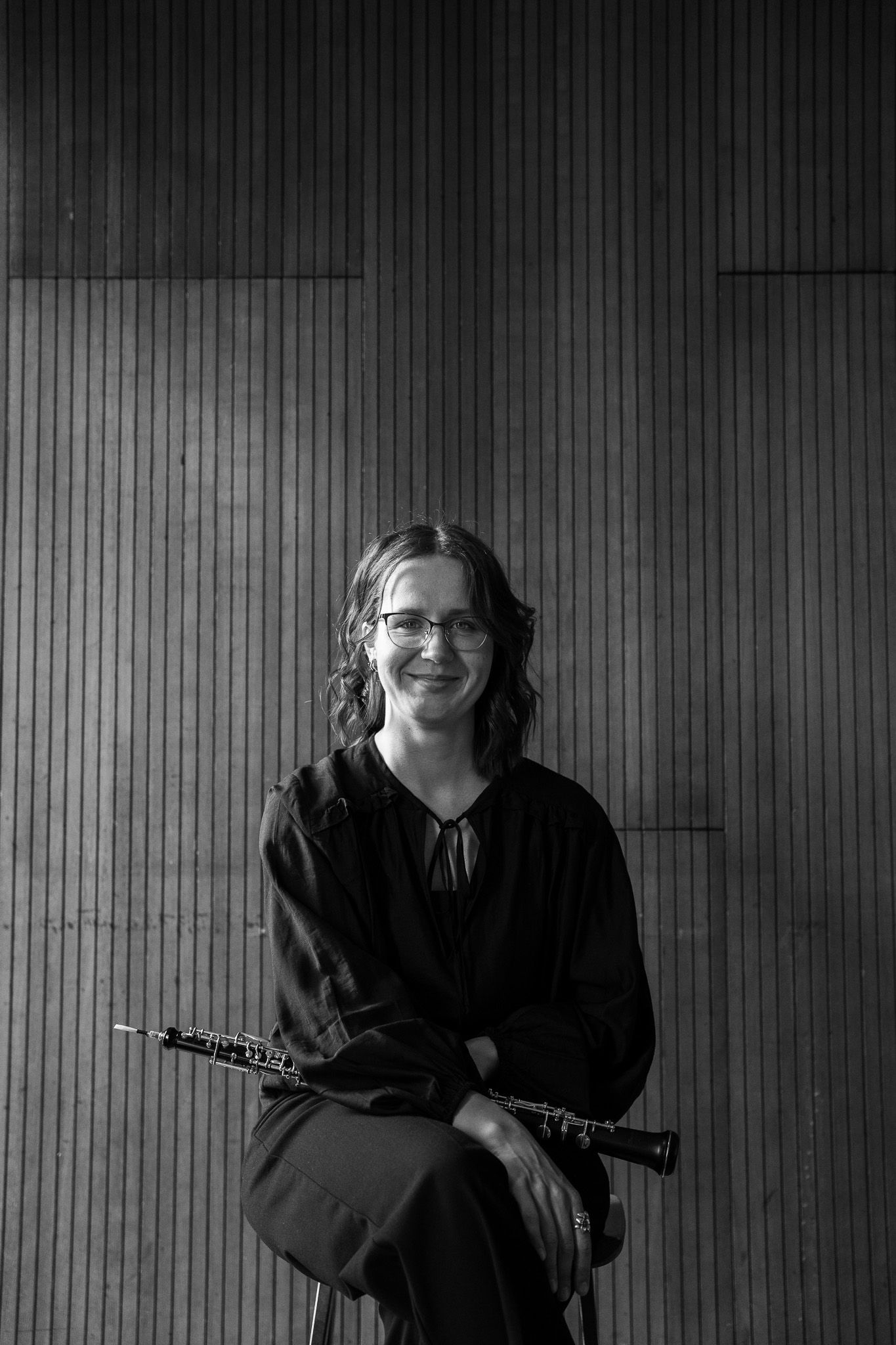 Smiling female oboist in black outfit seated with her oboe instrument in a black and white portrait photo.