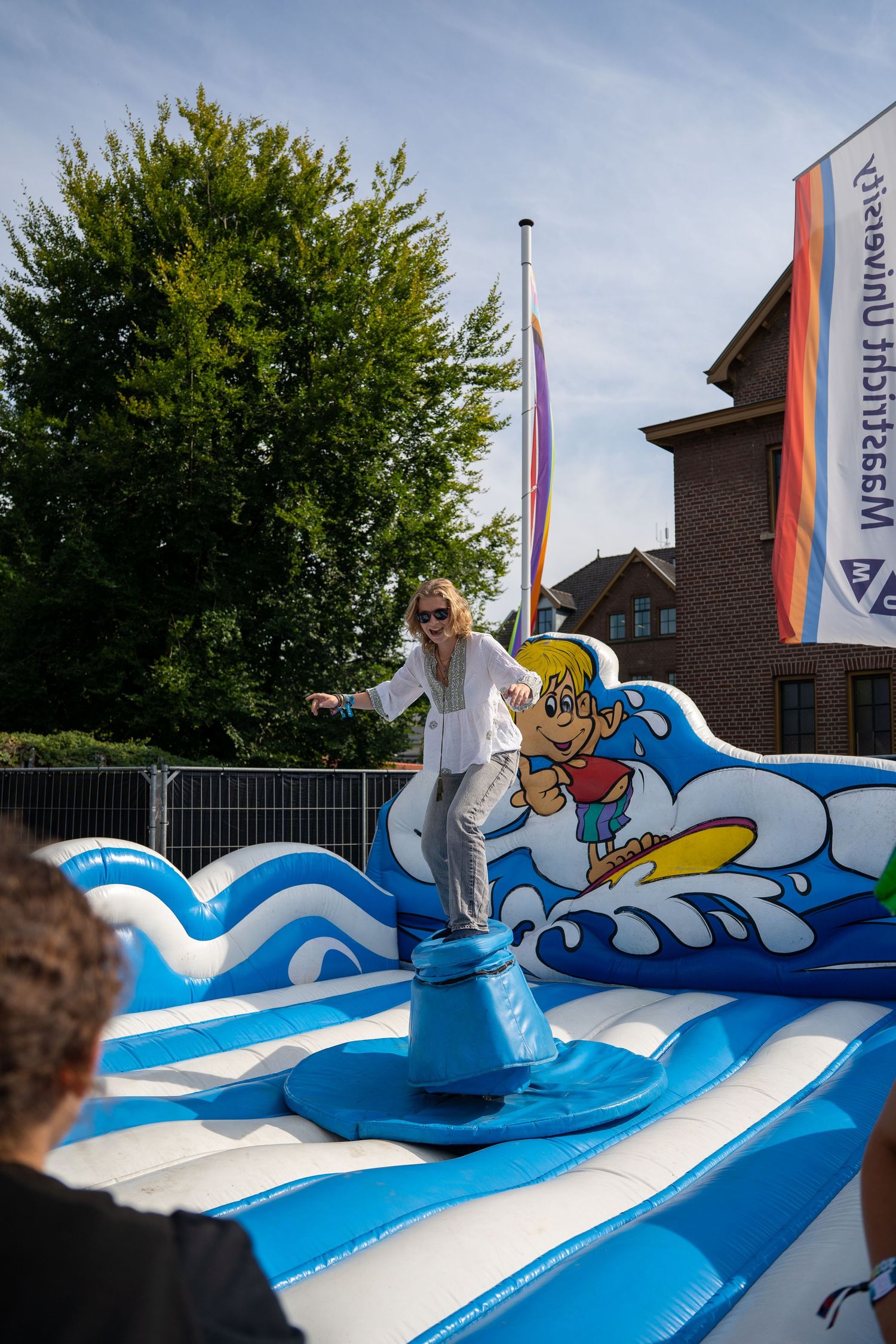 Blonde woman balancing on an inflatable mechanical surf ride at a sunny outdoor community festival.