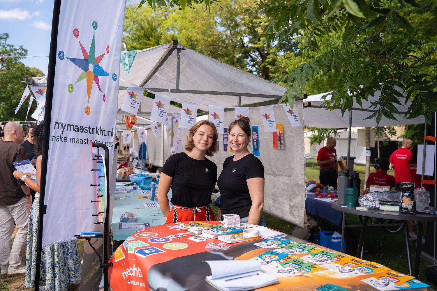 Two women in black shirts smile at a mymaastricht.nl information booth at an outdoor student fair.