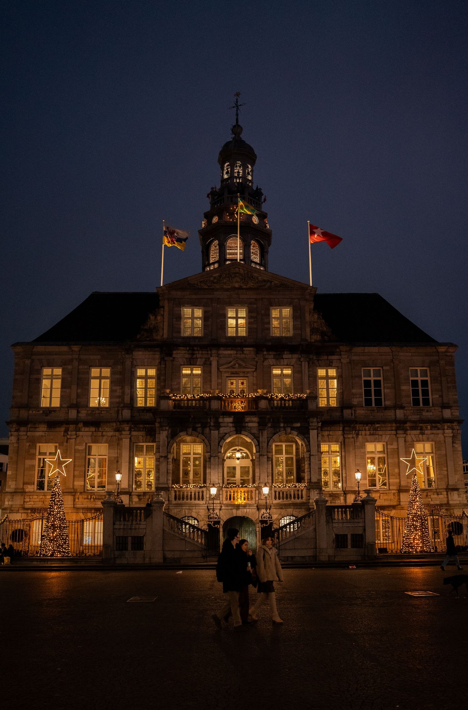 Historic illuminated town hall building decorated with Christmas trees and star lights at dusk.