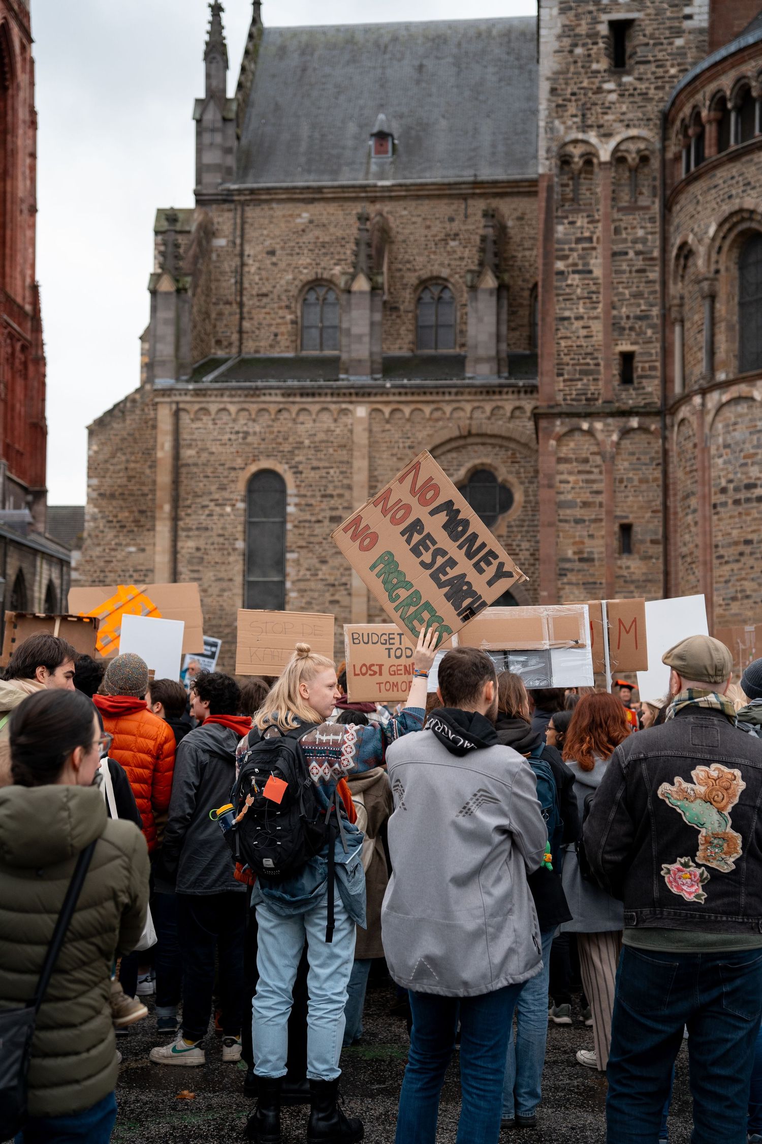 Protesters hold No Money No Research signs near historic brick church during education funding rally.