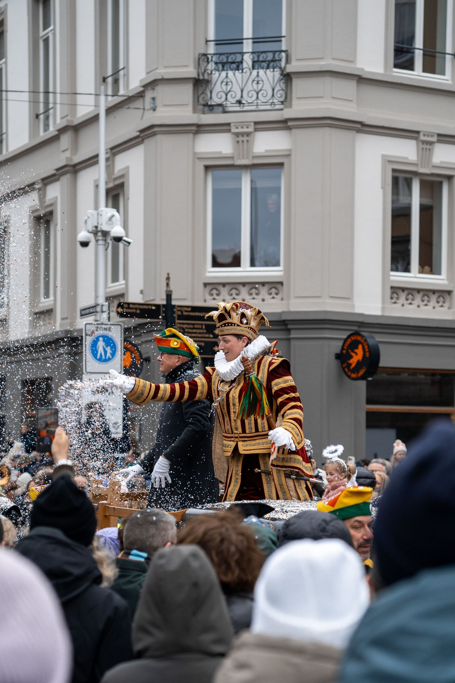 Carnival performer in royal costume tossing confetti from a float during a festive winter street parade.
