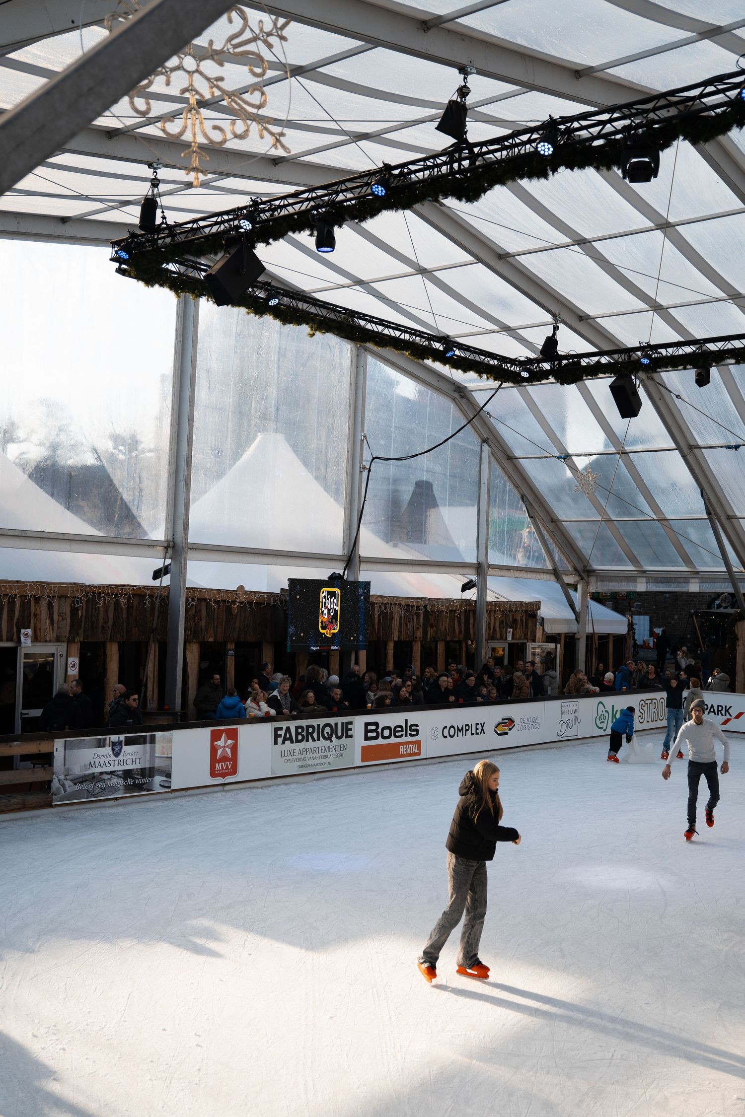 Indoor ice skating rink under a large transparent canopy with spectators and sponsor banners.