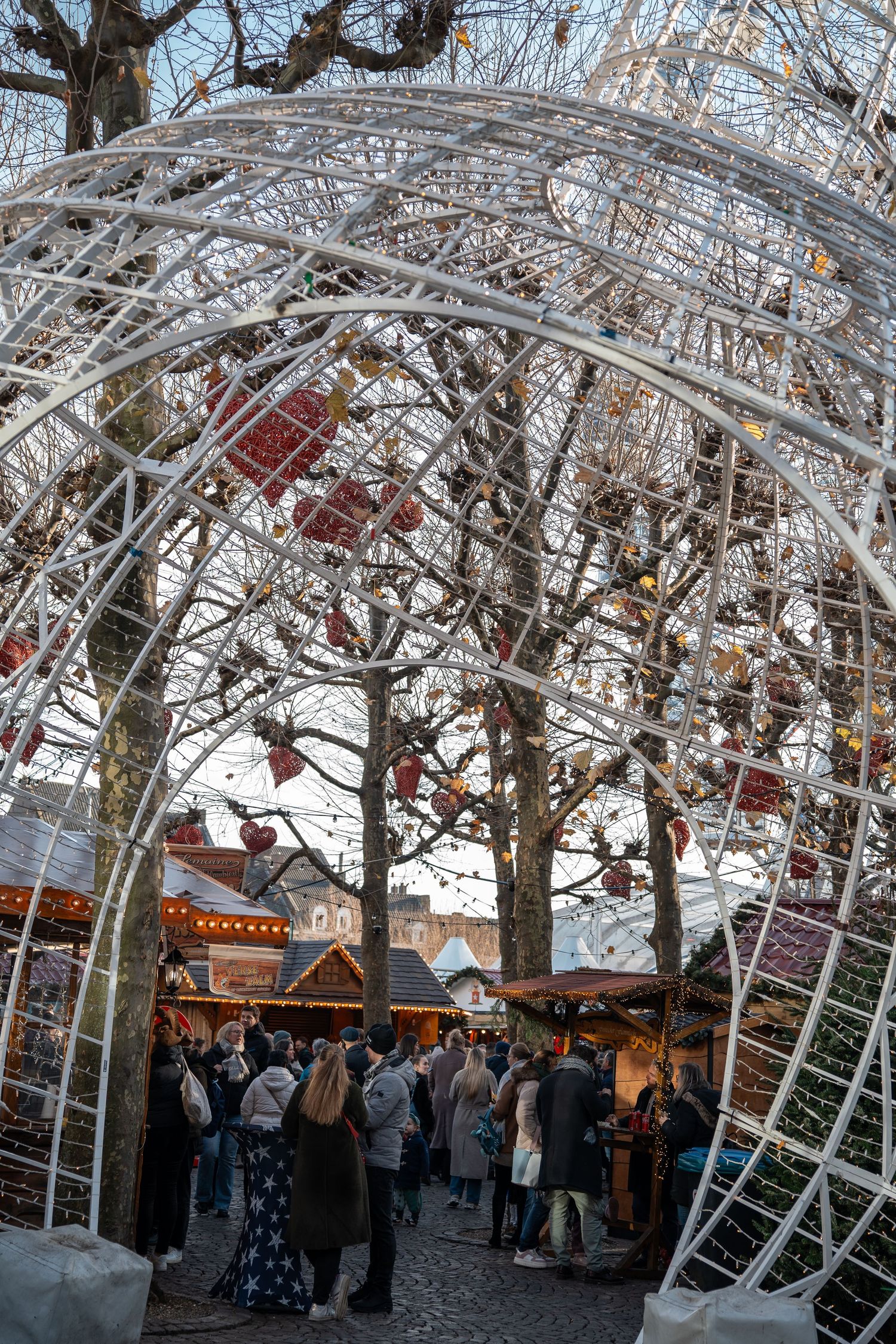 Crowds walking through a decorative white arch with red ornaments at a festive Christmas market.