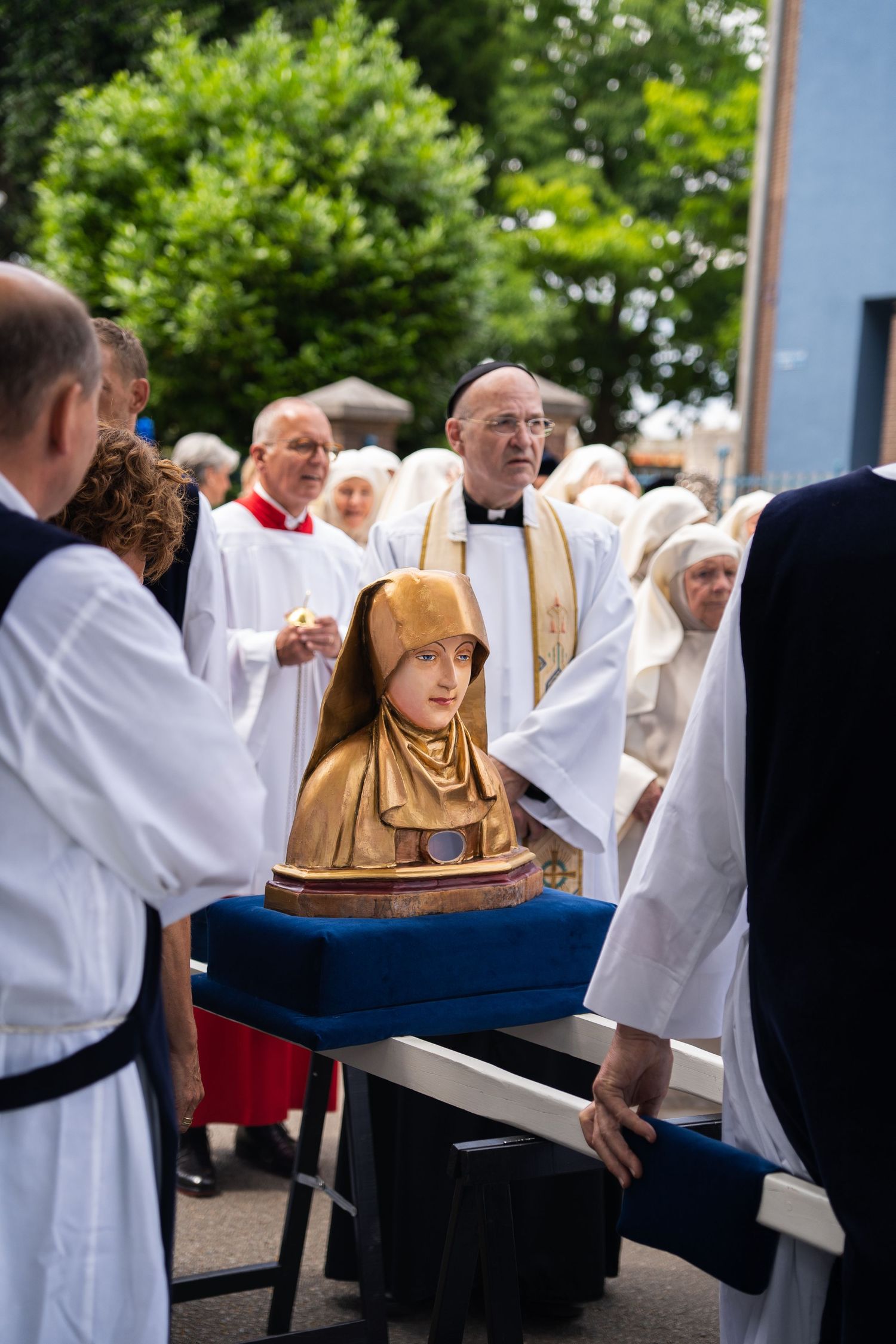 Priest oversees carved wooden saint bust carried on blue cushion in religious procession.