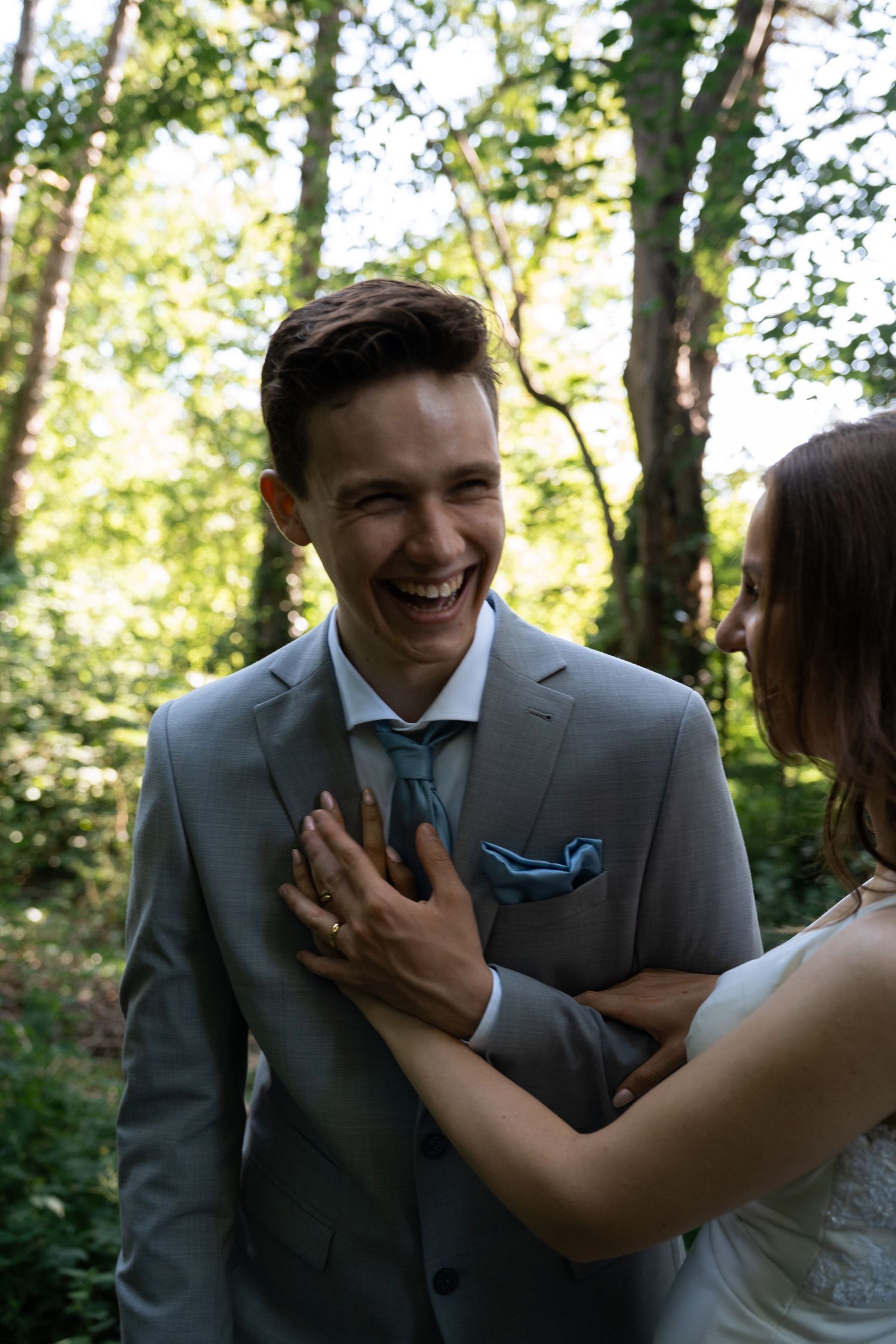 Laughing groom in gray suit with bride's hands on his chest in a sunlit forest setting.