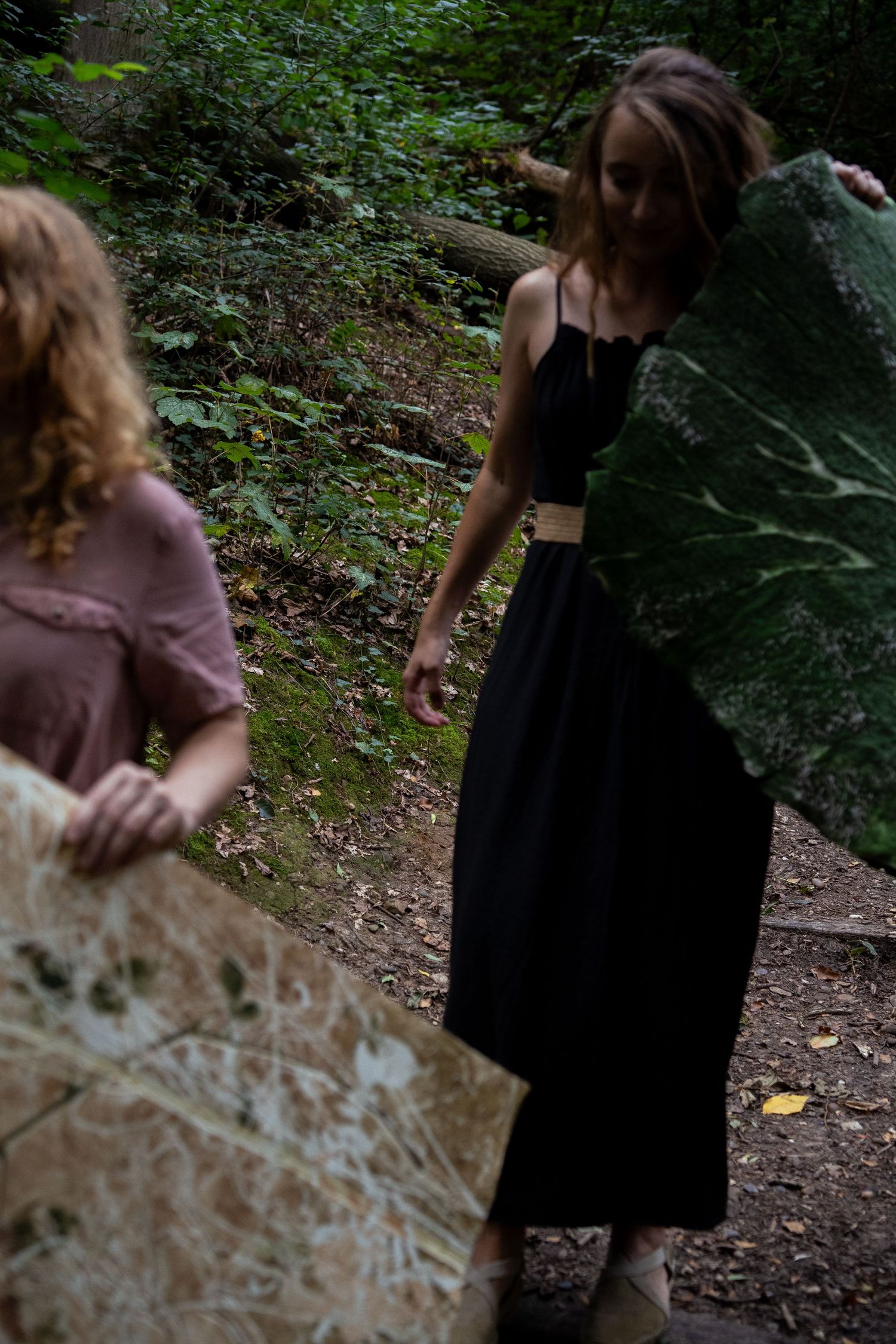 Two women carrying large nature-printed artworks along a woodland forest path.