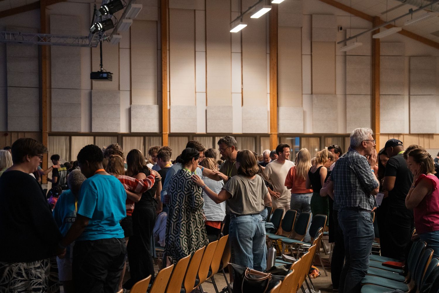 People standing and praying together in rows inside a warmly lit wooden community hall.