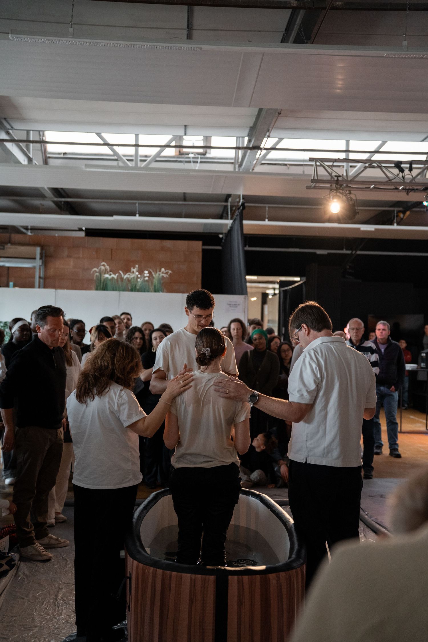 Group of people in white shirts gathered around a baptismal font during a church baptism ceremony indoors.