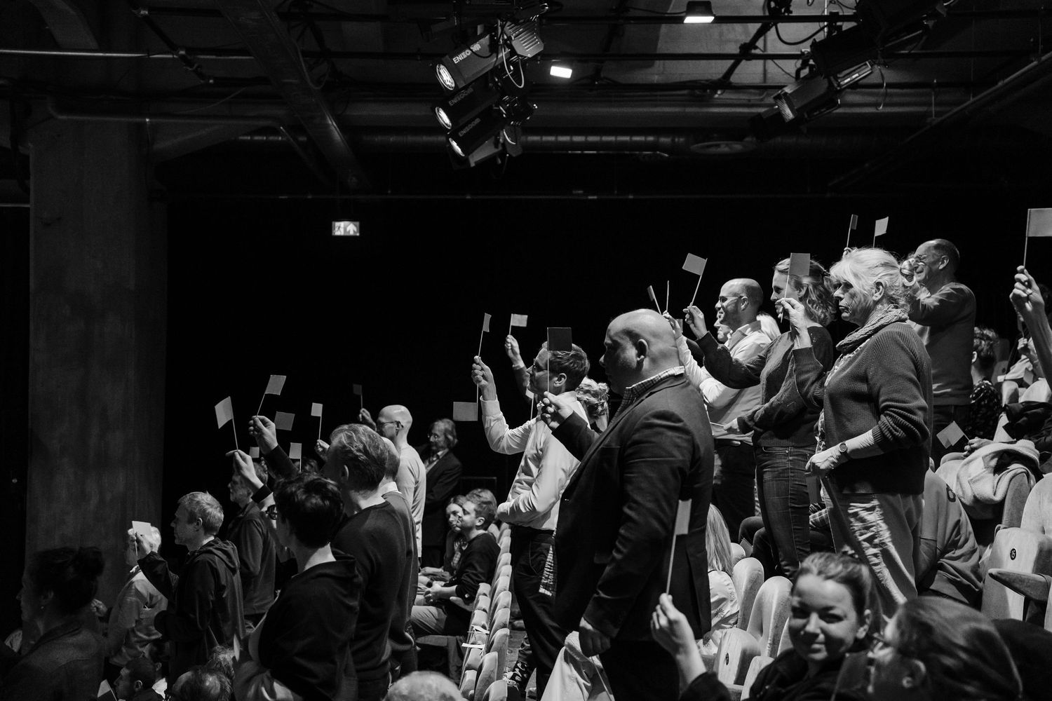 Black and white image of engaged audience holding up phones at an indoor theatrical performance.