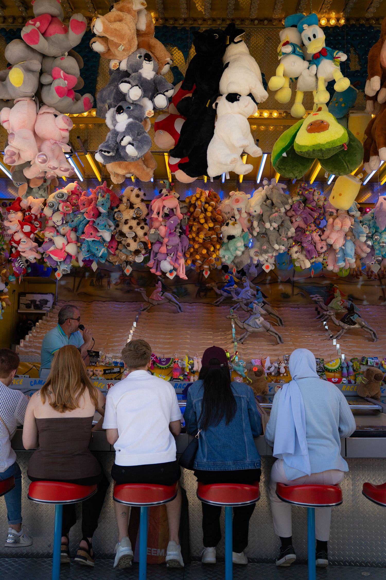 People sitting on red stools at a carnival game booth filled with colorful stuffed animal prizes hanging overhead.