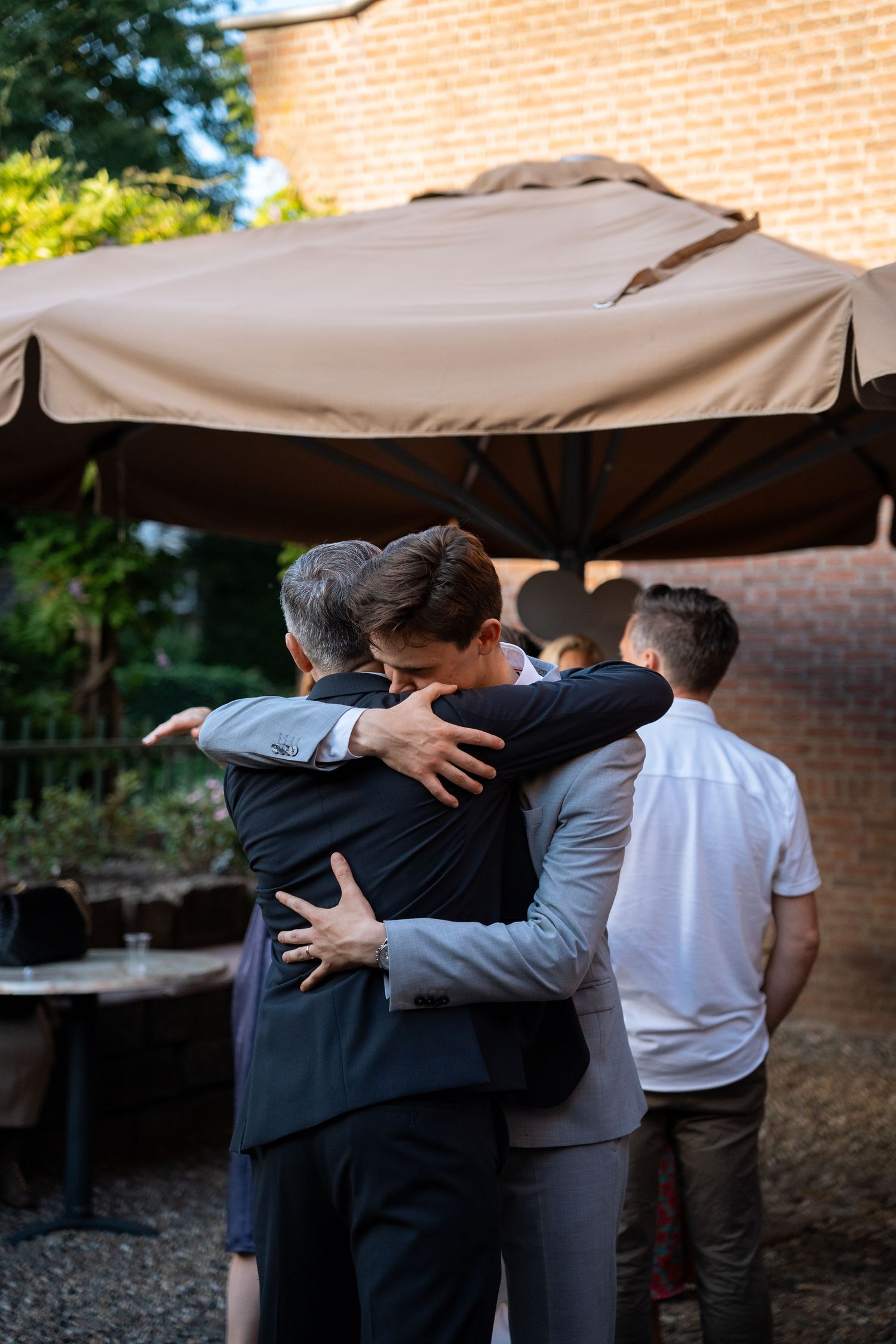 Two men share a heartfelt embrace at an outdoor wedding reception under a large beige patio umbrella.
