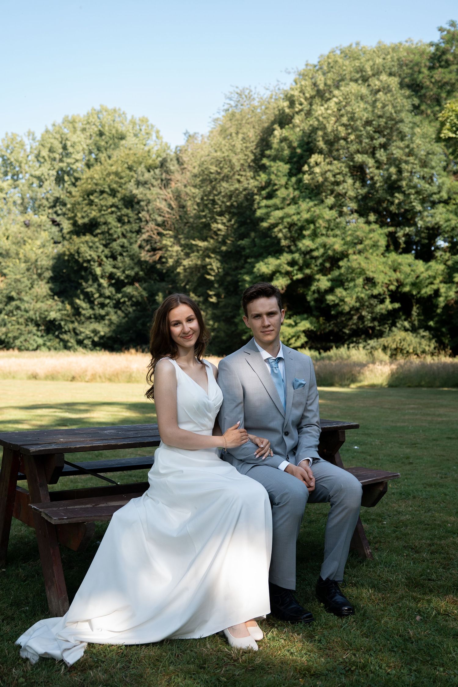 Bride and groom sitting together on a wooden bench in a sunny green park.
