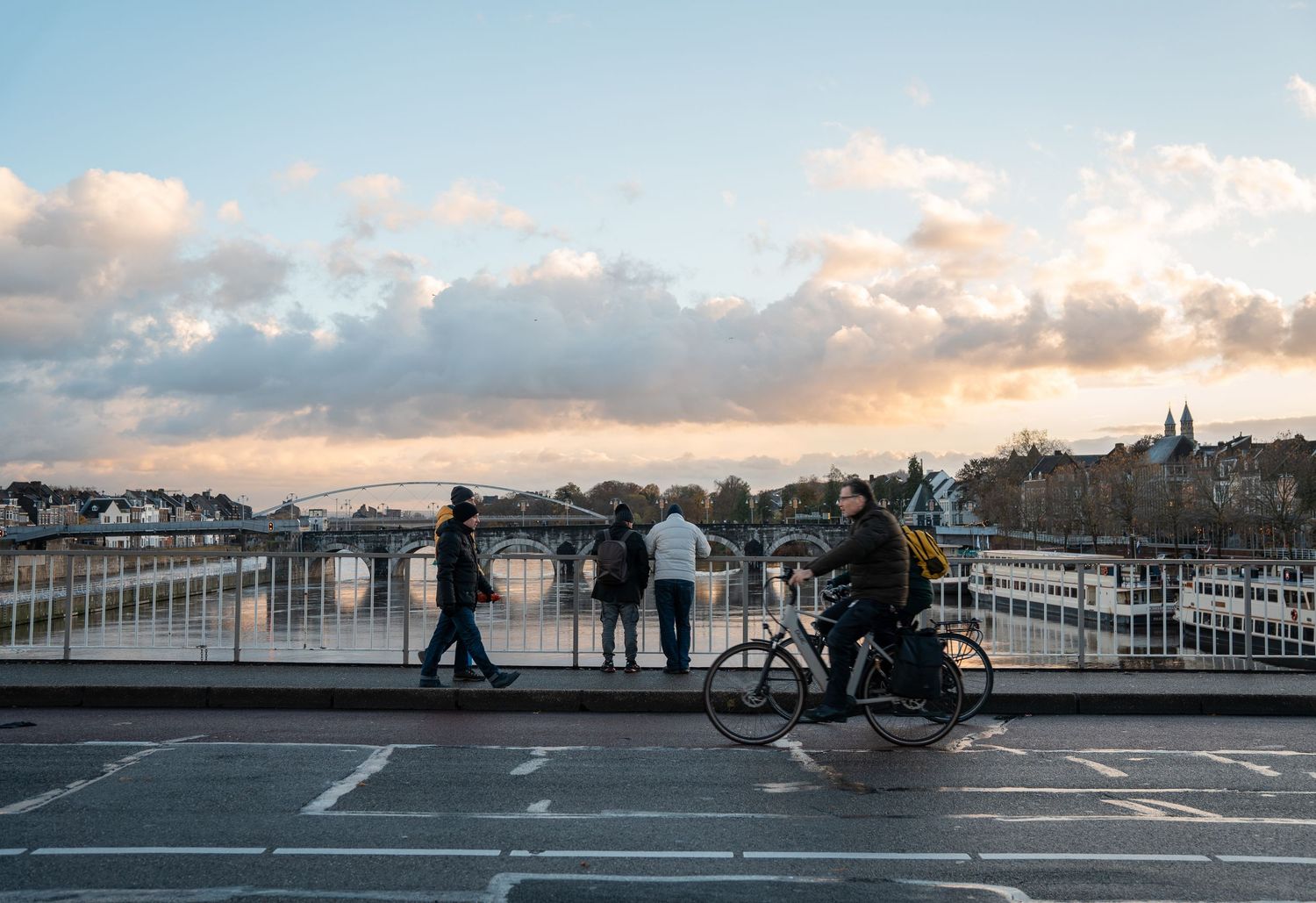 People walking and cycling on a bridge at sunset with a river and city skyline in the background.