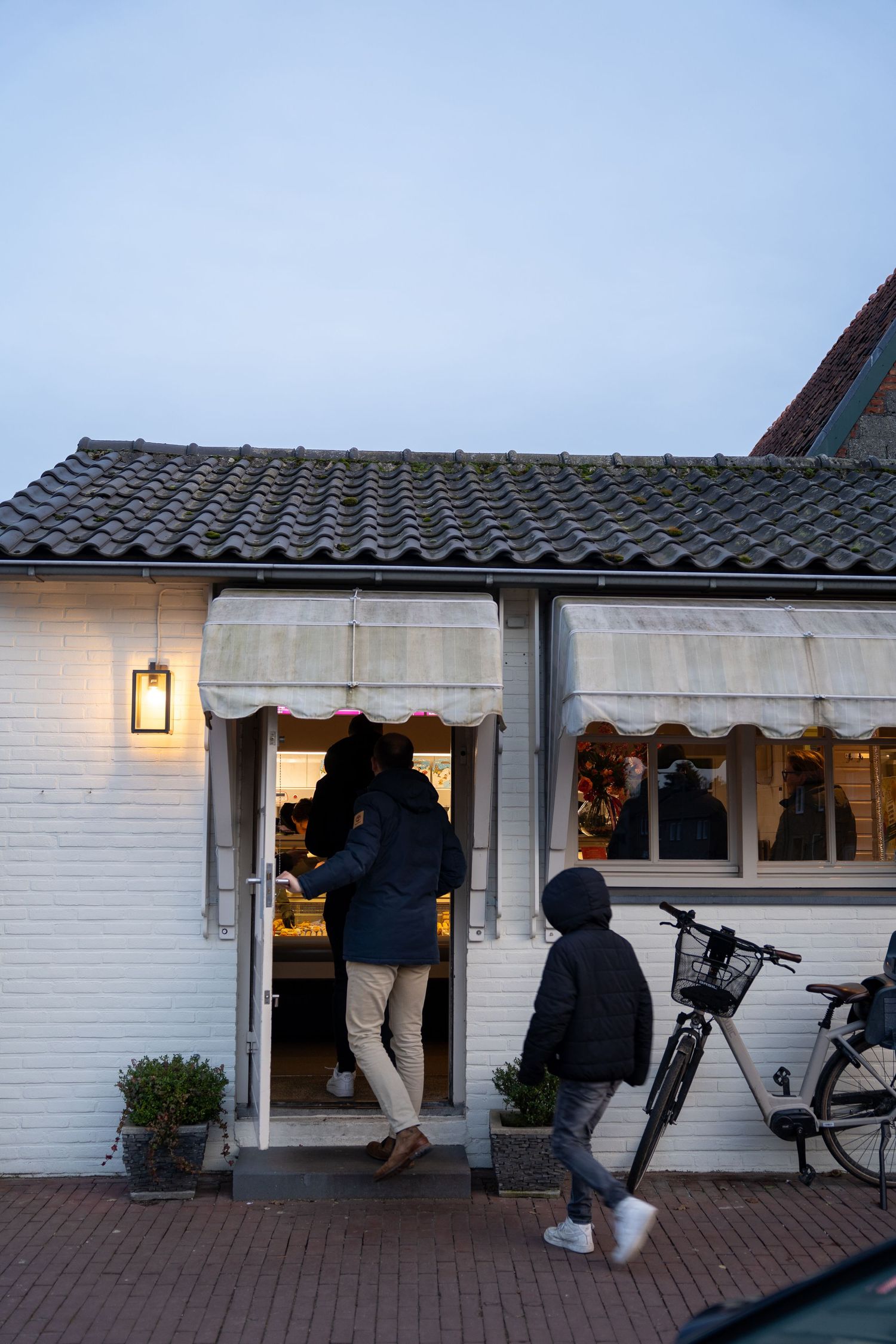 Person entering a cozy white brick shop at dusk with a child and bicycle outside, warm lantern light glowing above.