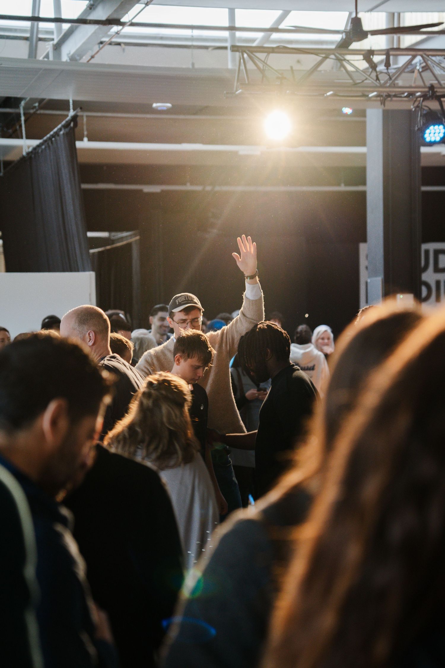Large crowd at an indoor worship event with one person raising their hand under a bright spotlight.