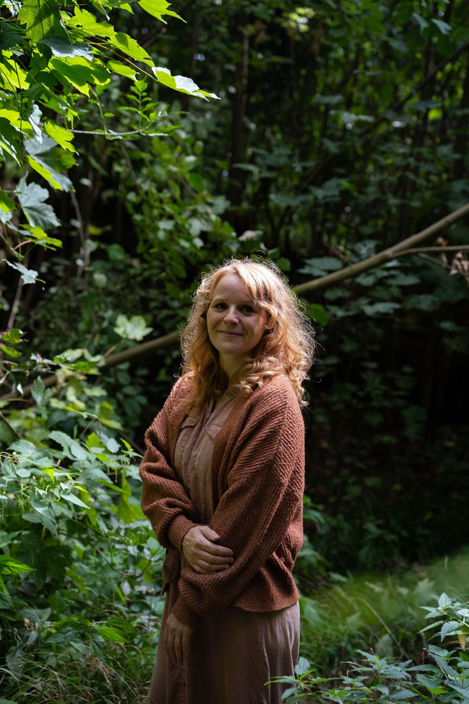 Smiling woman with curly red hair standing in a lush green forest.