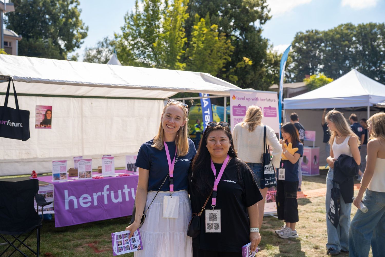 Two women wearing lanyards pose together at an outdoor herfuture career fair booth on a sunny day.