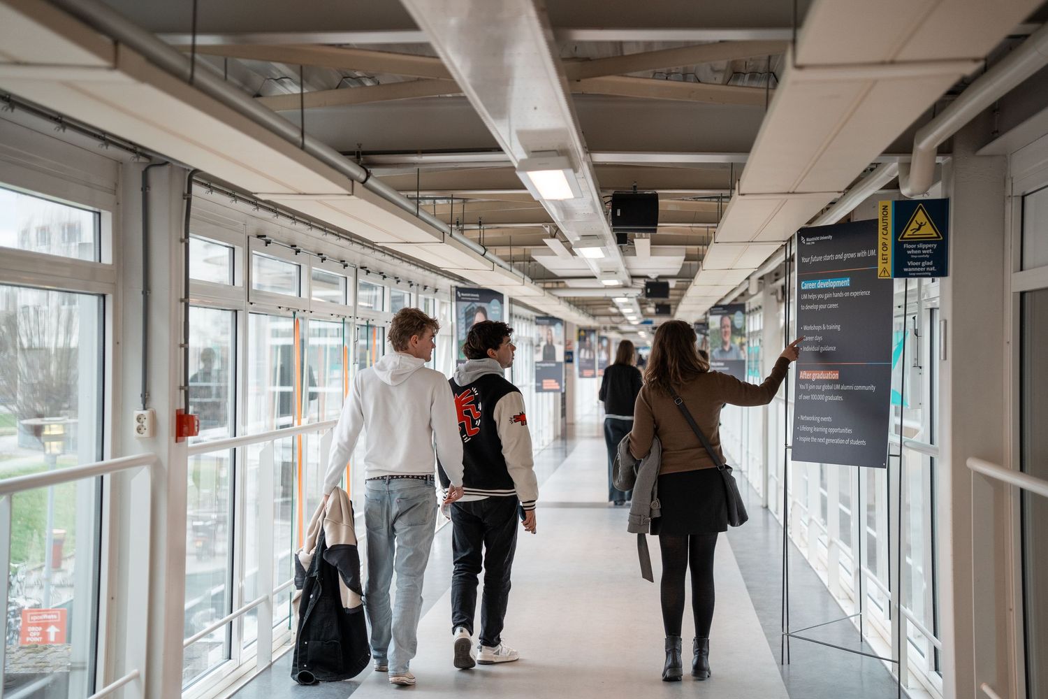 Students walking through modern glass corridor bridge at university campus building.