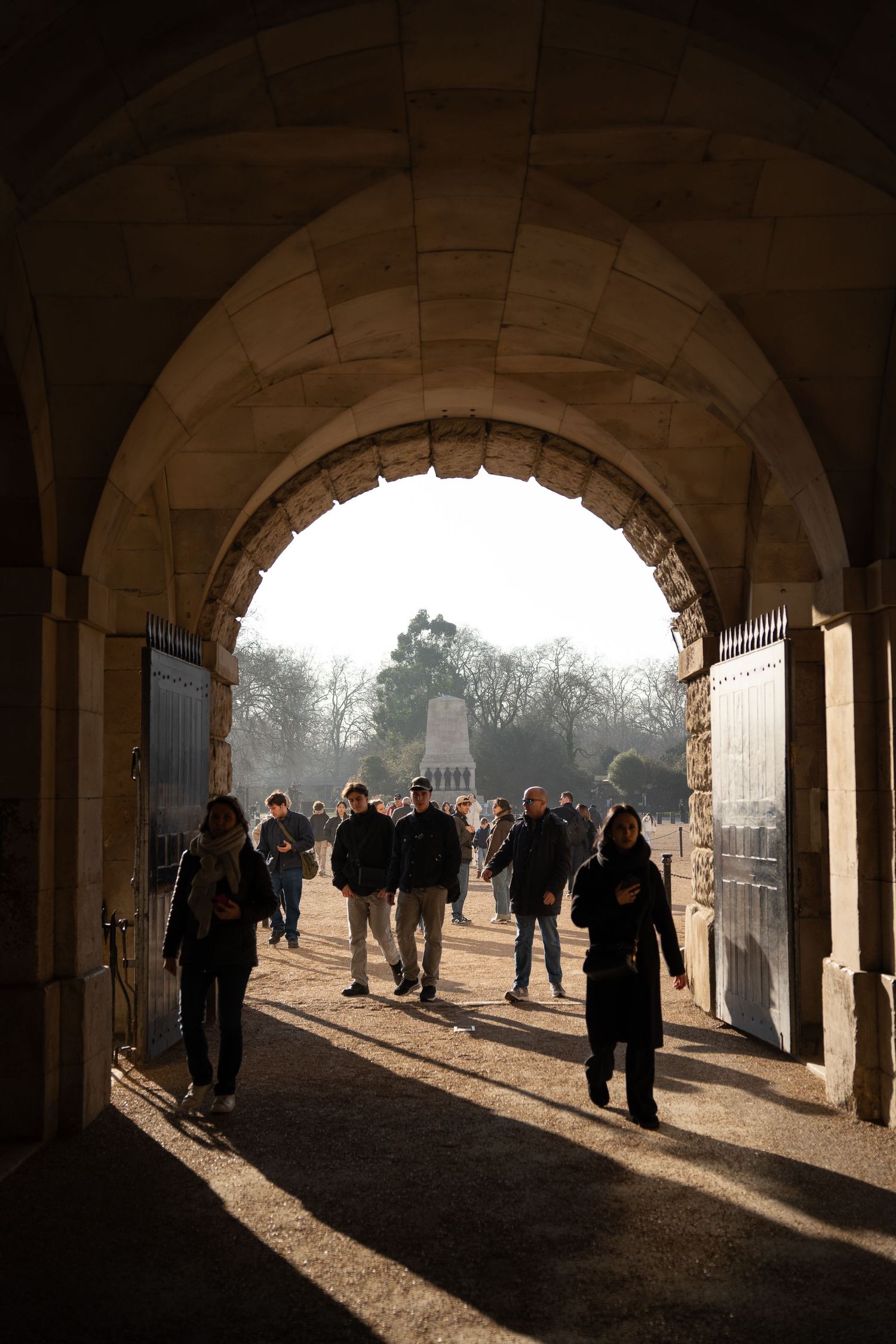 Silhouetted visitors walking through a grand stone archway into a sunlit park with a monument visible beyond.