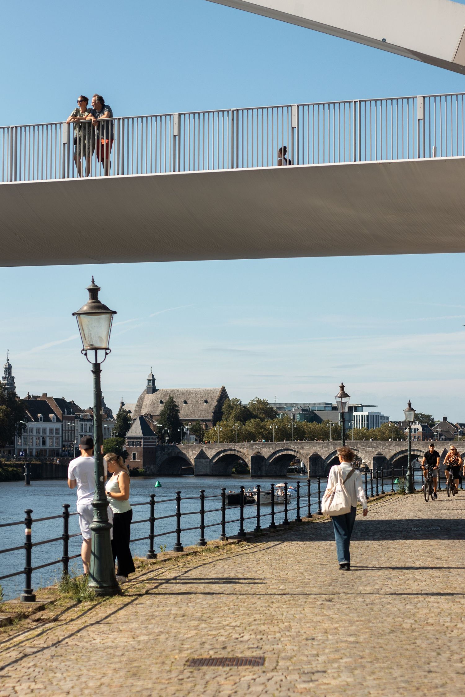 People walking along a riverside promenade in Maastricht with a modern footbridge and historic Sint Servaasbrug in the background.
