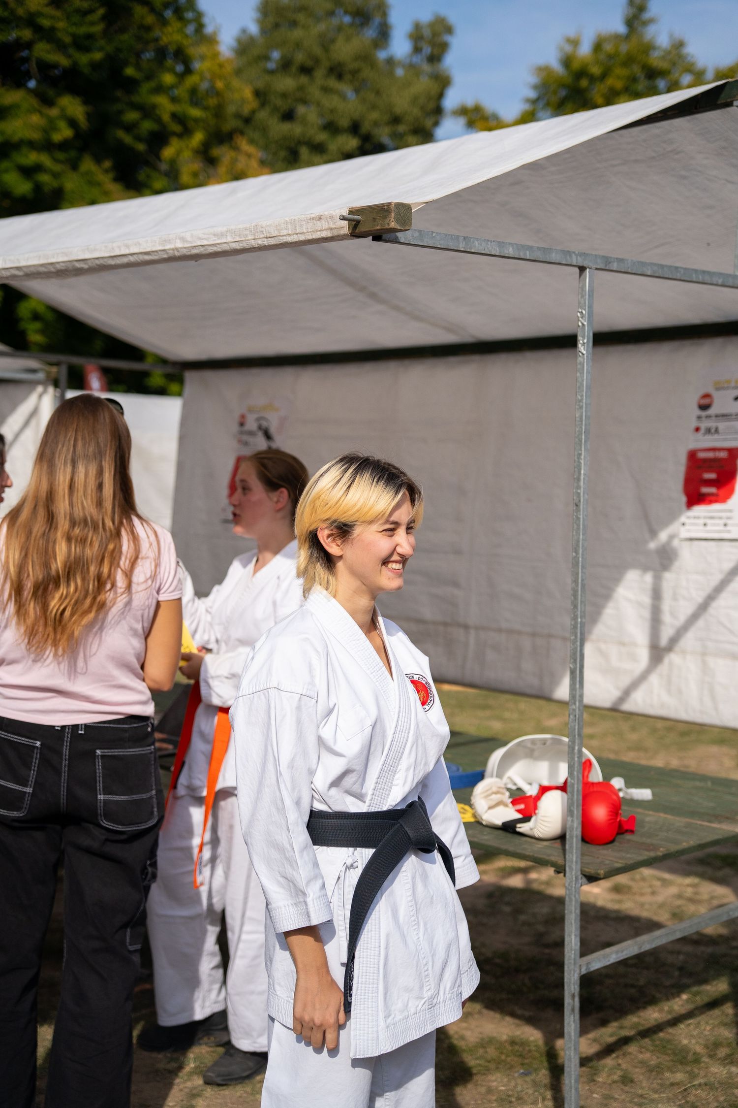 Smiling woman in white karate gi with black belt standing under a canopy at an outdoor martial arts event.