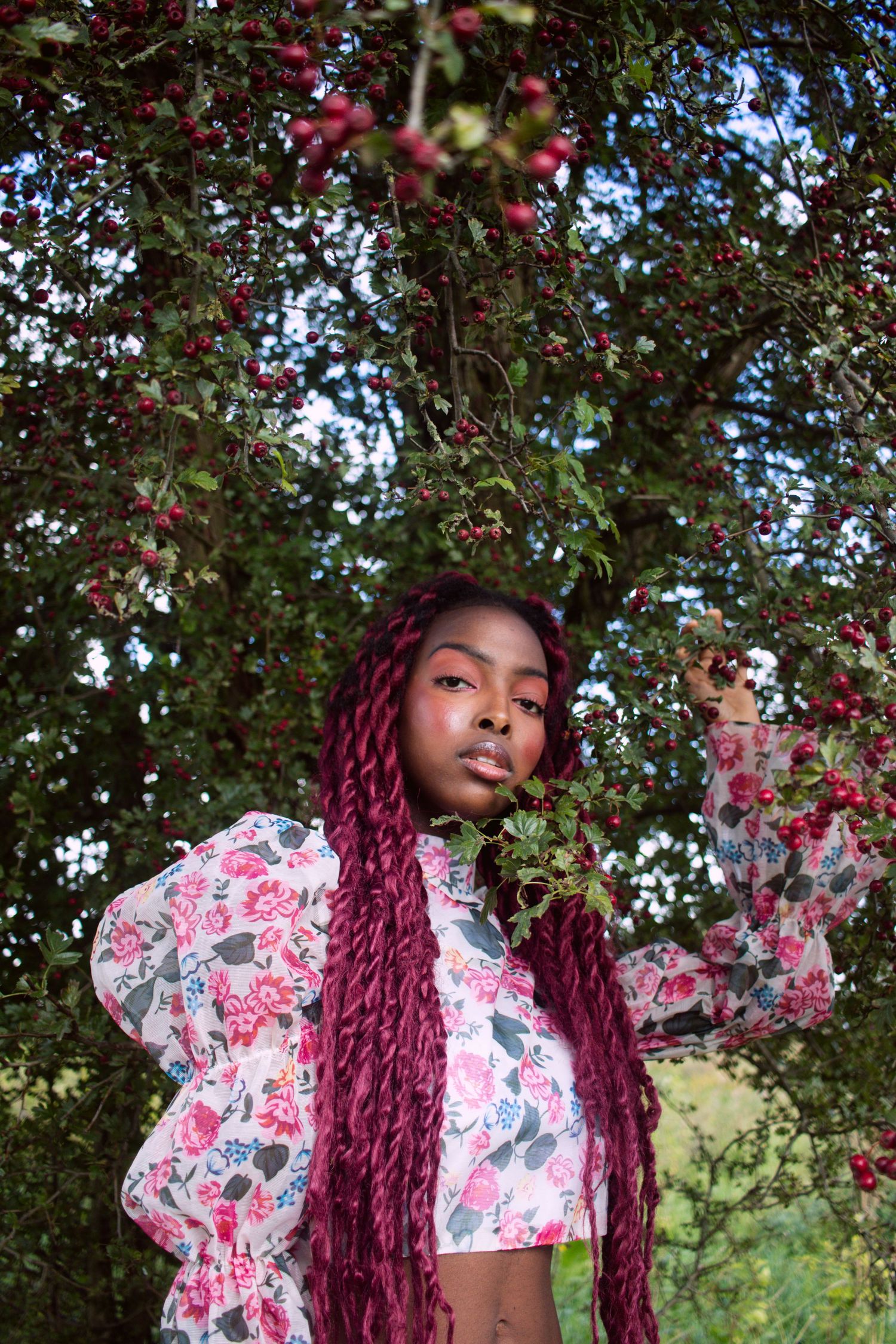 Young Black woman with long red braids in floral crop top poses under berry-laden tree branches.