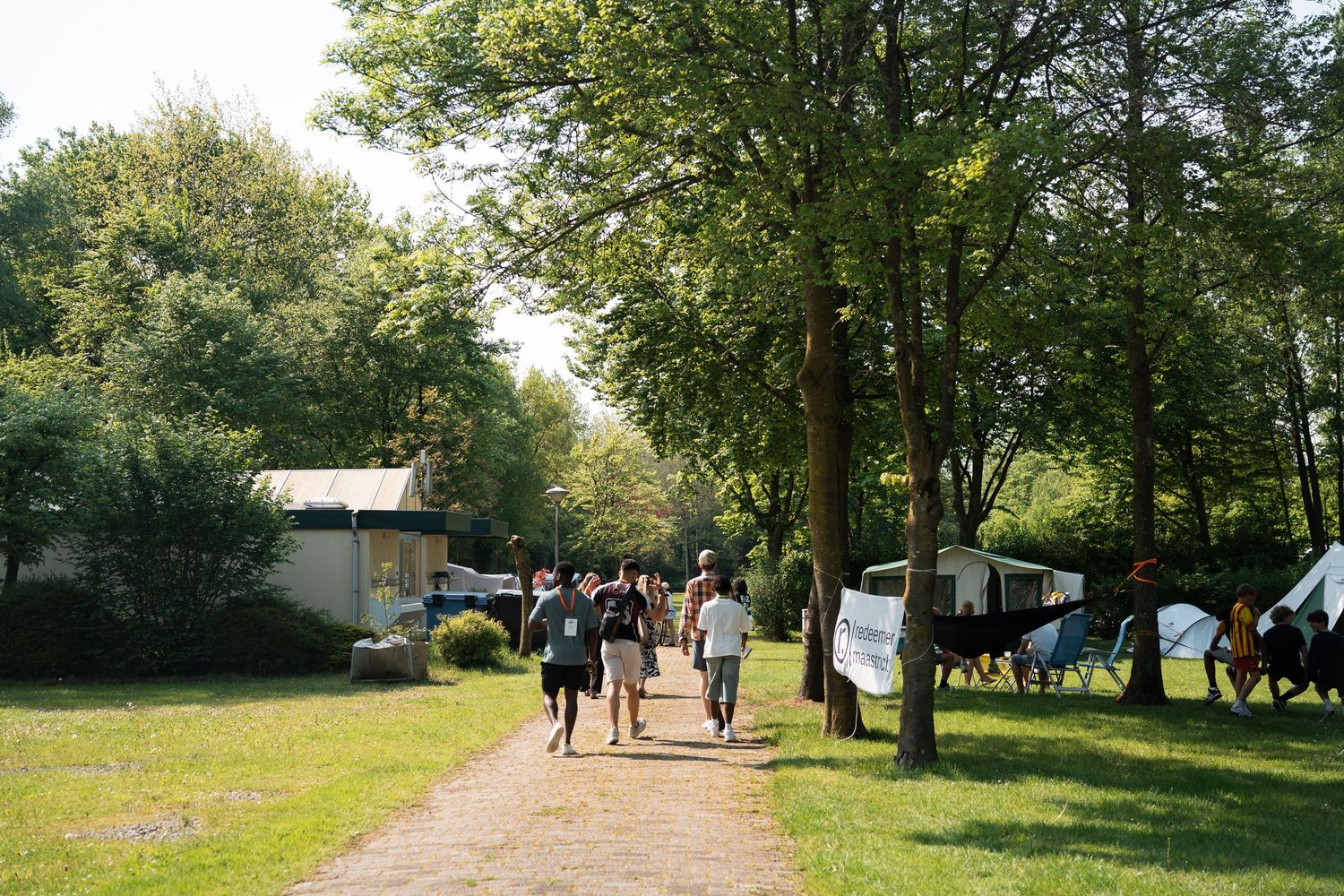 People walking along a tree-lined path past tents and cabins at an outdoor summer camp festival.