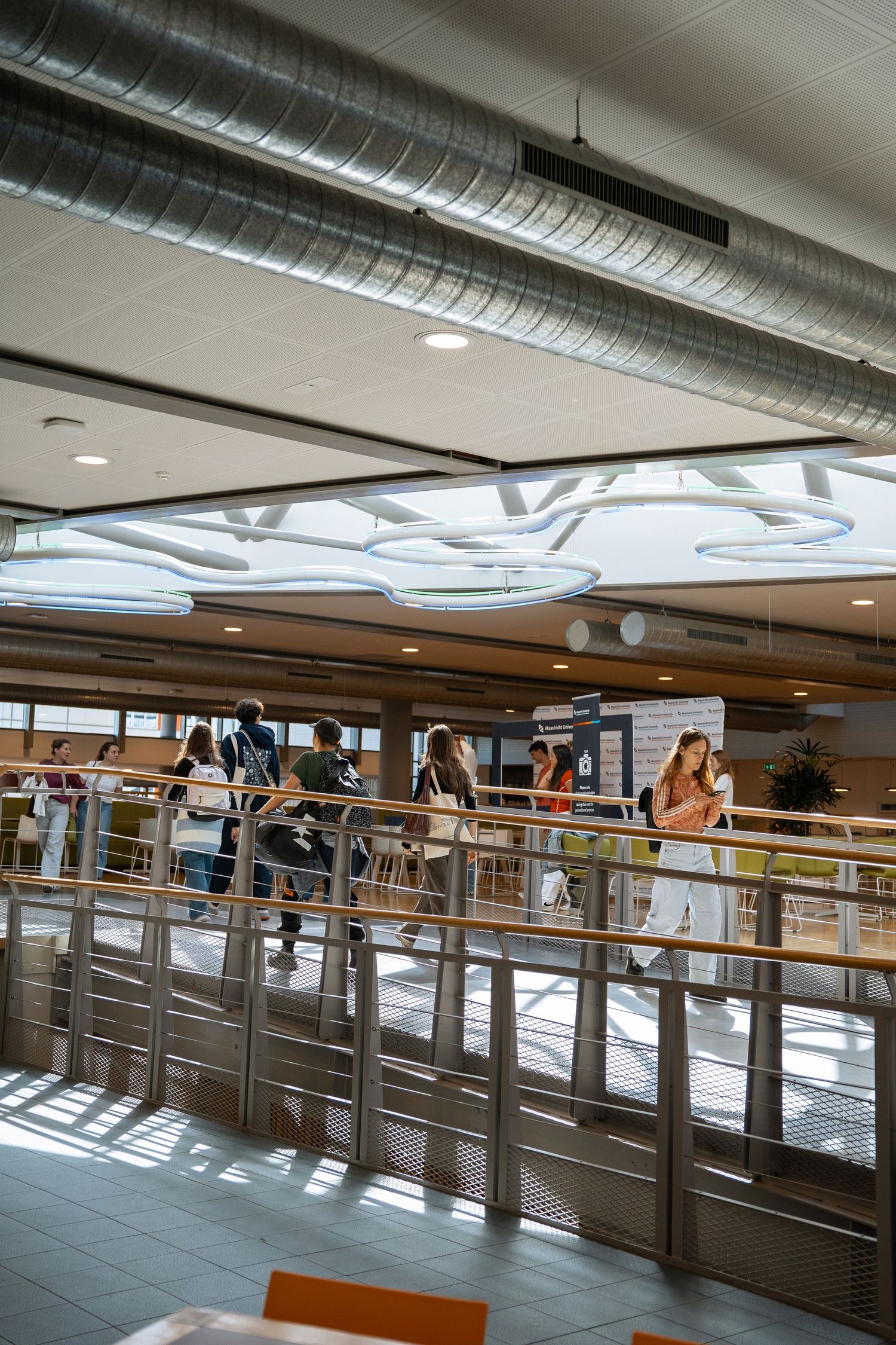 Students walking along a modern university mezzanine with metal railings and decorative ceiling lights.
