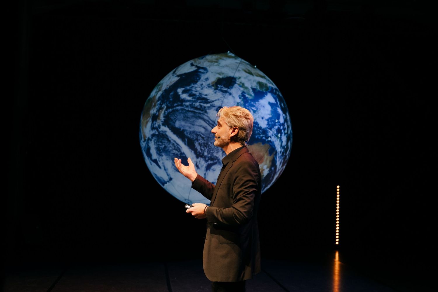 Speaker presenting on stage with large illuminated Earth globe projection in dark theater setting.