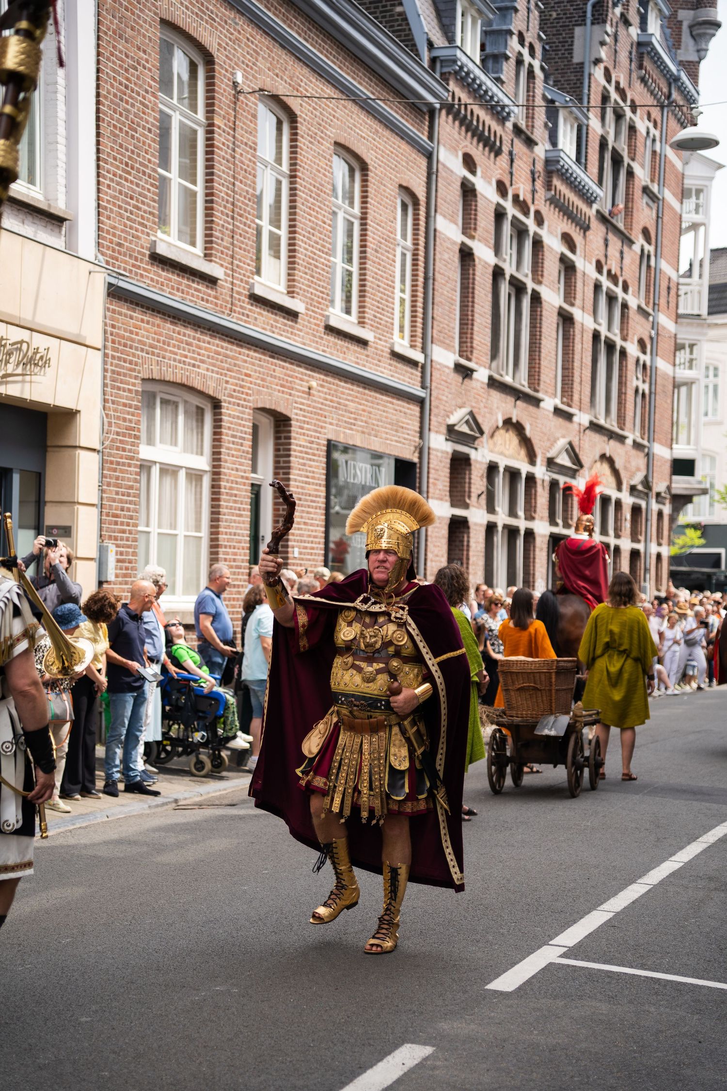 Performer in golden Roman armor leads a street parade past brick buildings and spectators.