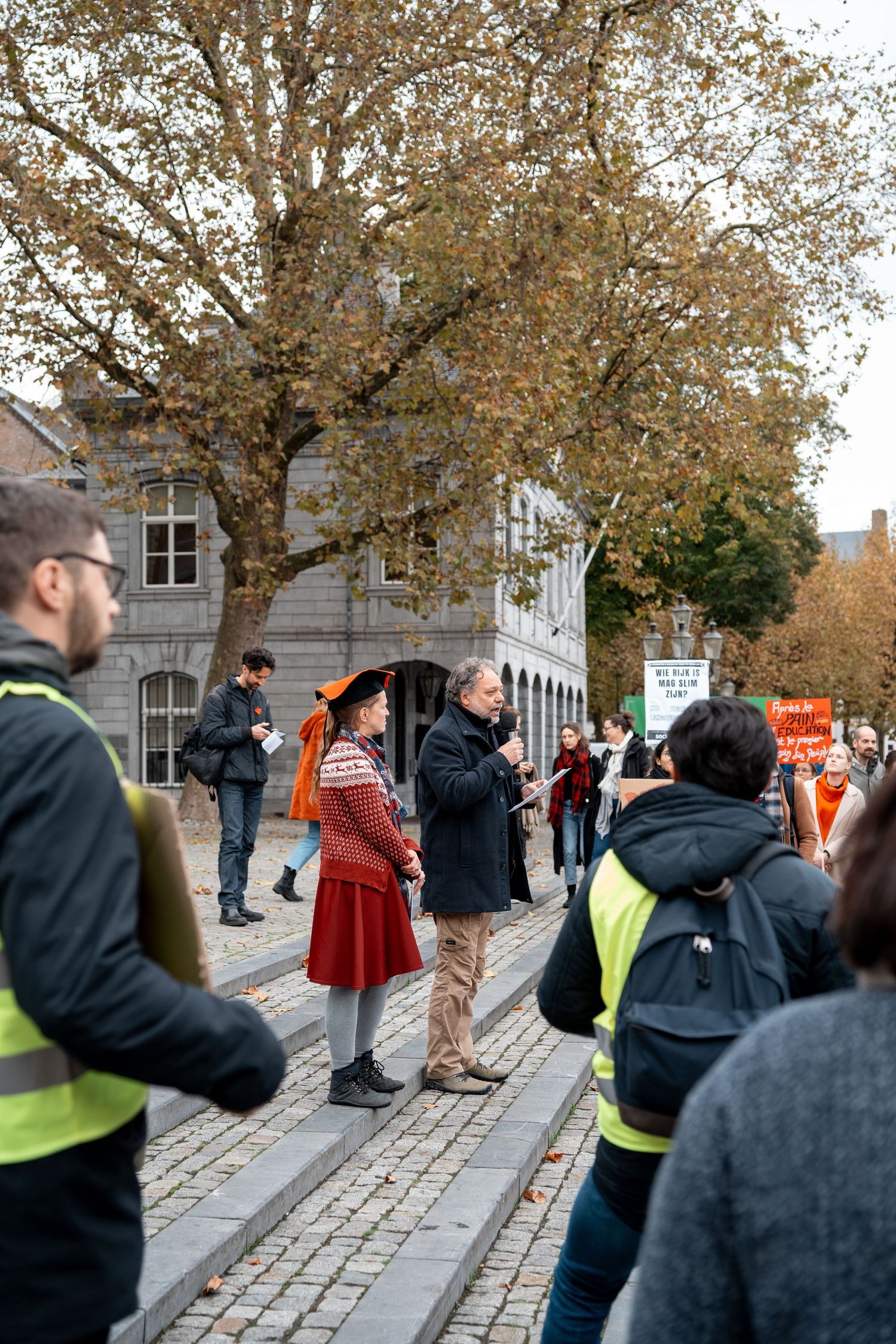 Woman in red dress and man converse among yellow-vested marshals at outdoor protest event.