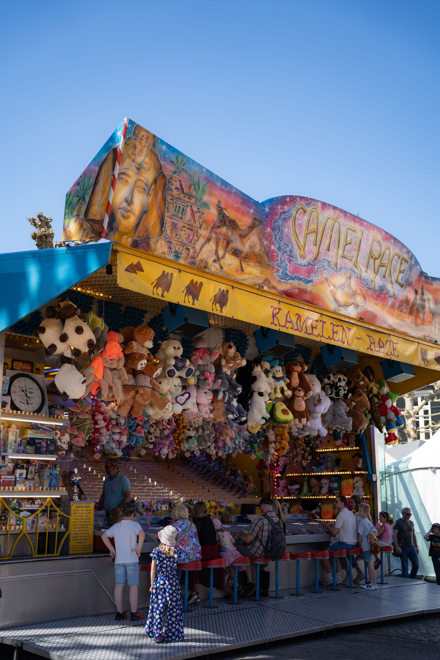 Visitors browse a Camel Race carnival game booth decorated with colorful stuffed animal prizes at a fairground.