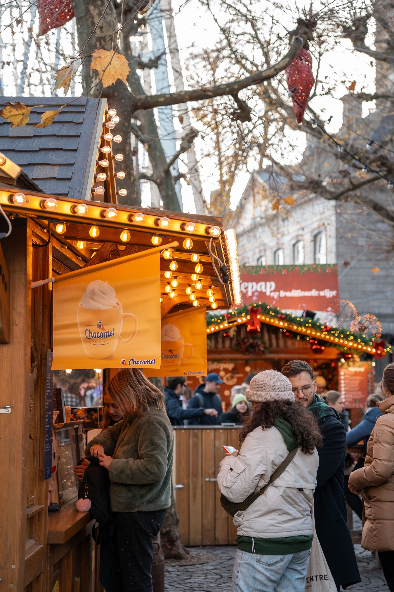Visitors browsing a warmly lit Christmas market stall selling hot chocolate drinks outdoors.
