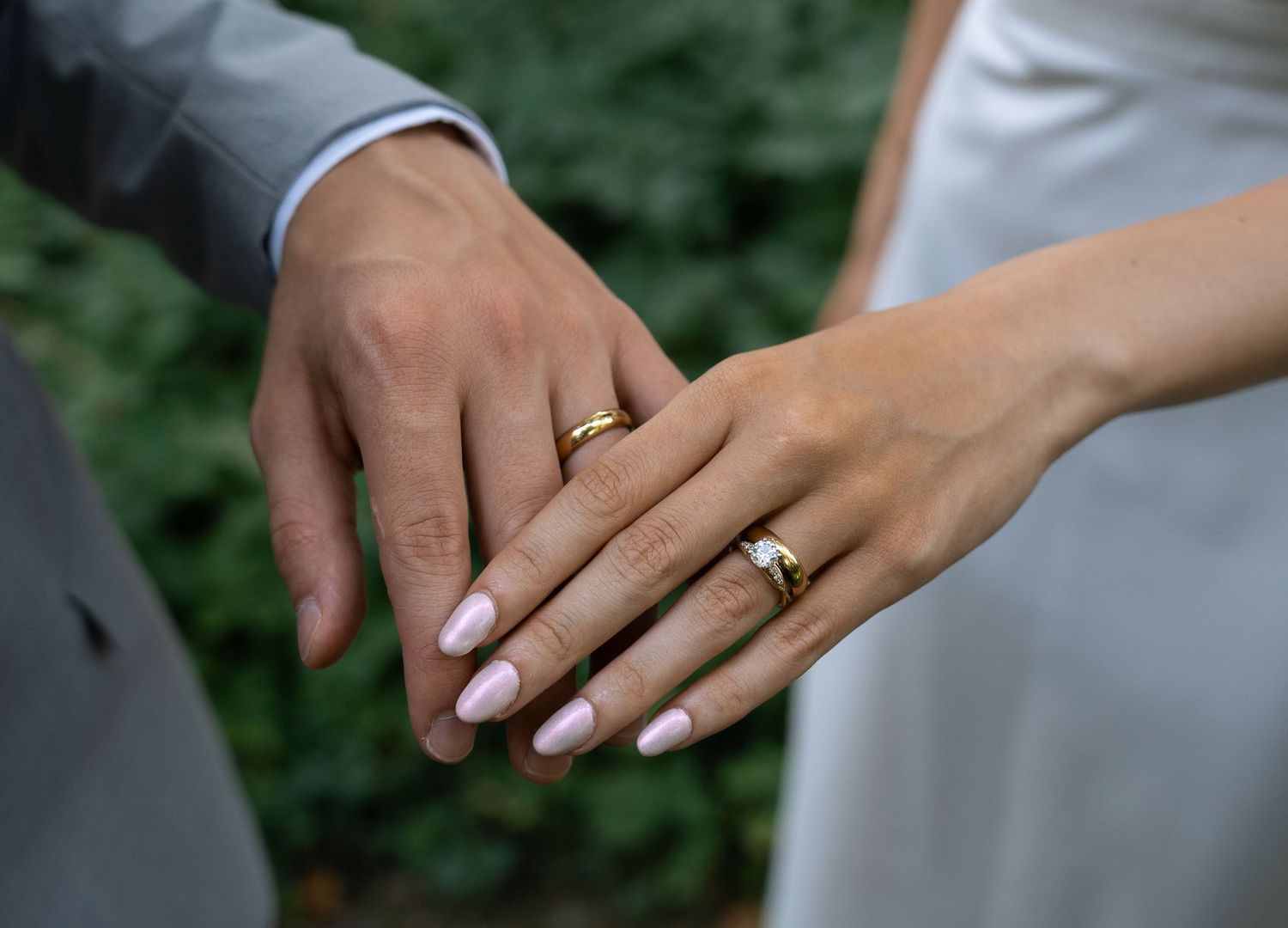 Close-up of newlyweds holding hands displaying matching gold wedding rings with pink manicured nails.