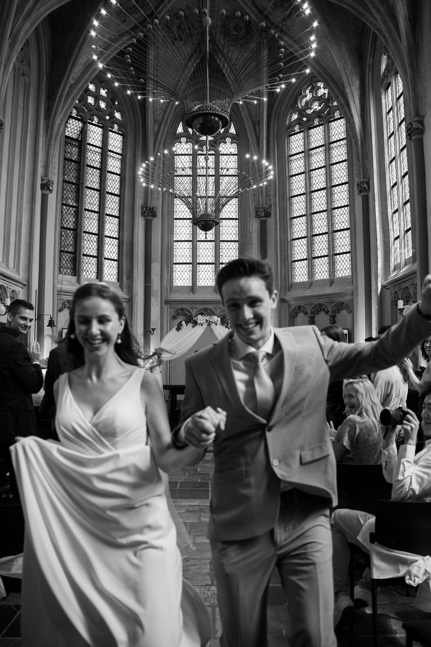 Black and white photo of joyful newlyweds walking down the aisle in a grand Gothic church after their wedding ceremony.