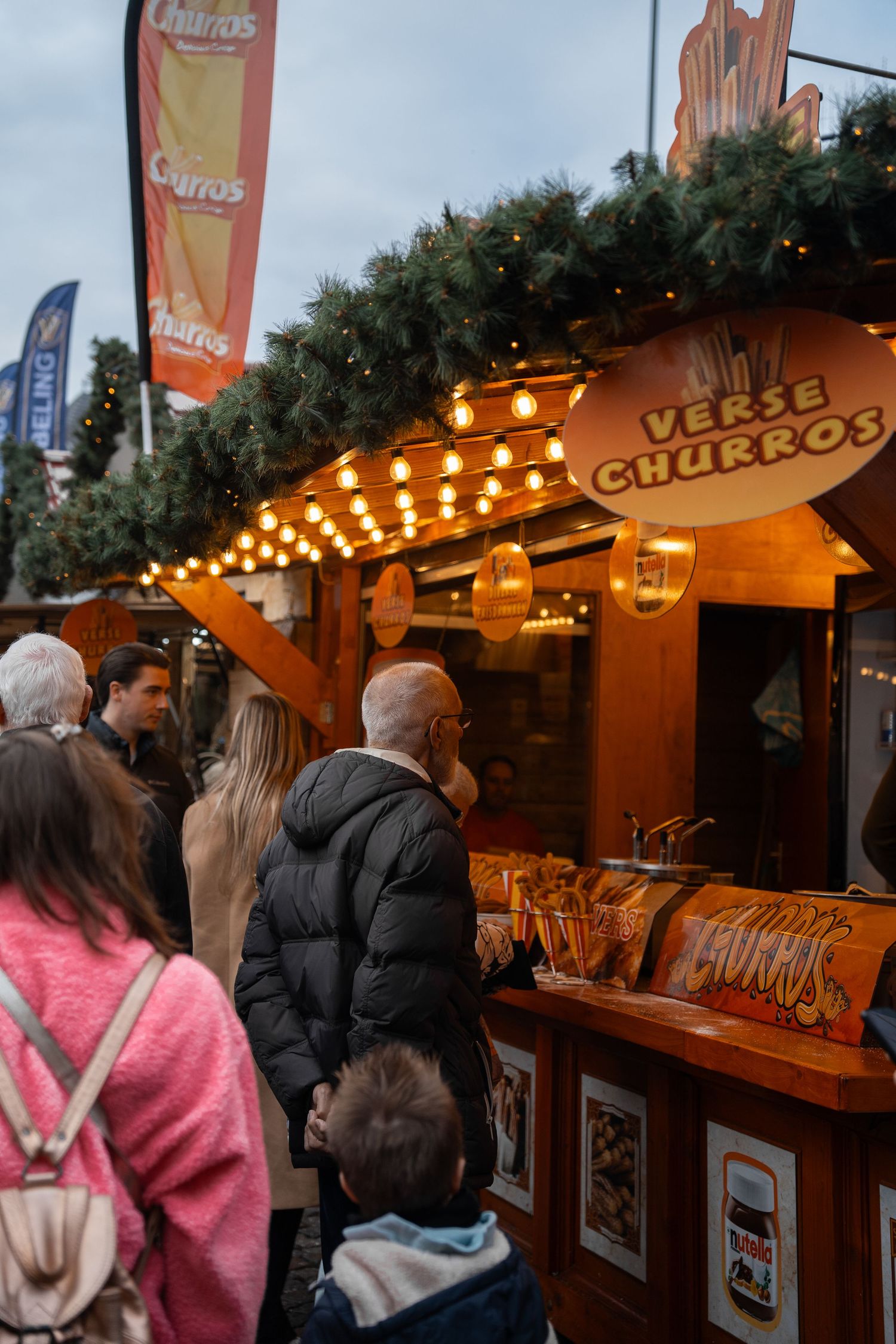 Customers queuing at a festive wooden churros stall decorated with Christmas garlands and warm lights.