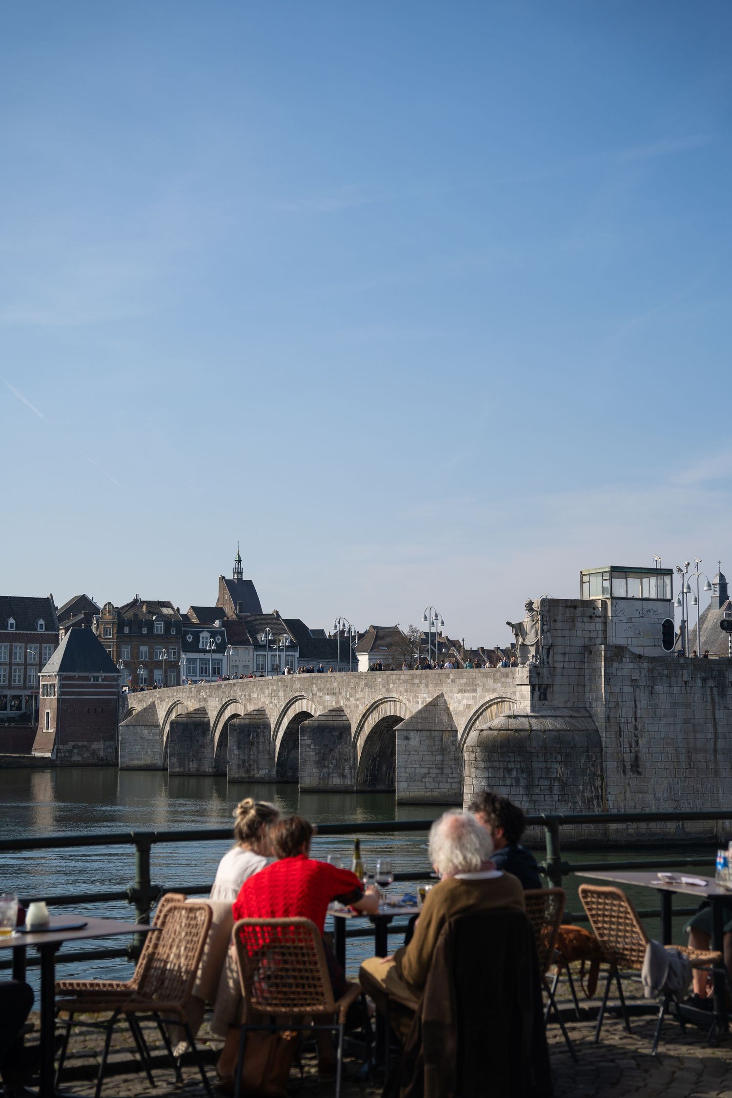 People dining at a riverside terrace overlooking the historic Sint Servaas Bridge in Maastricht on a sunny day.
