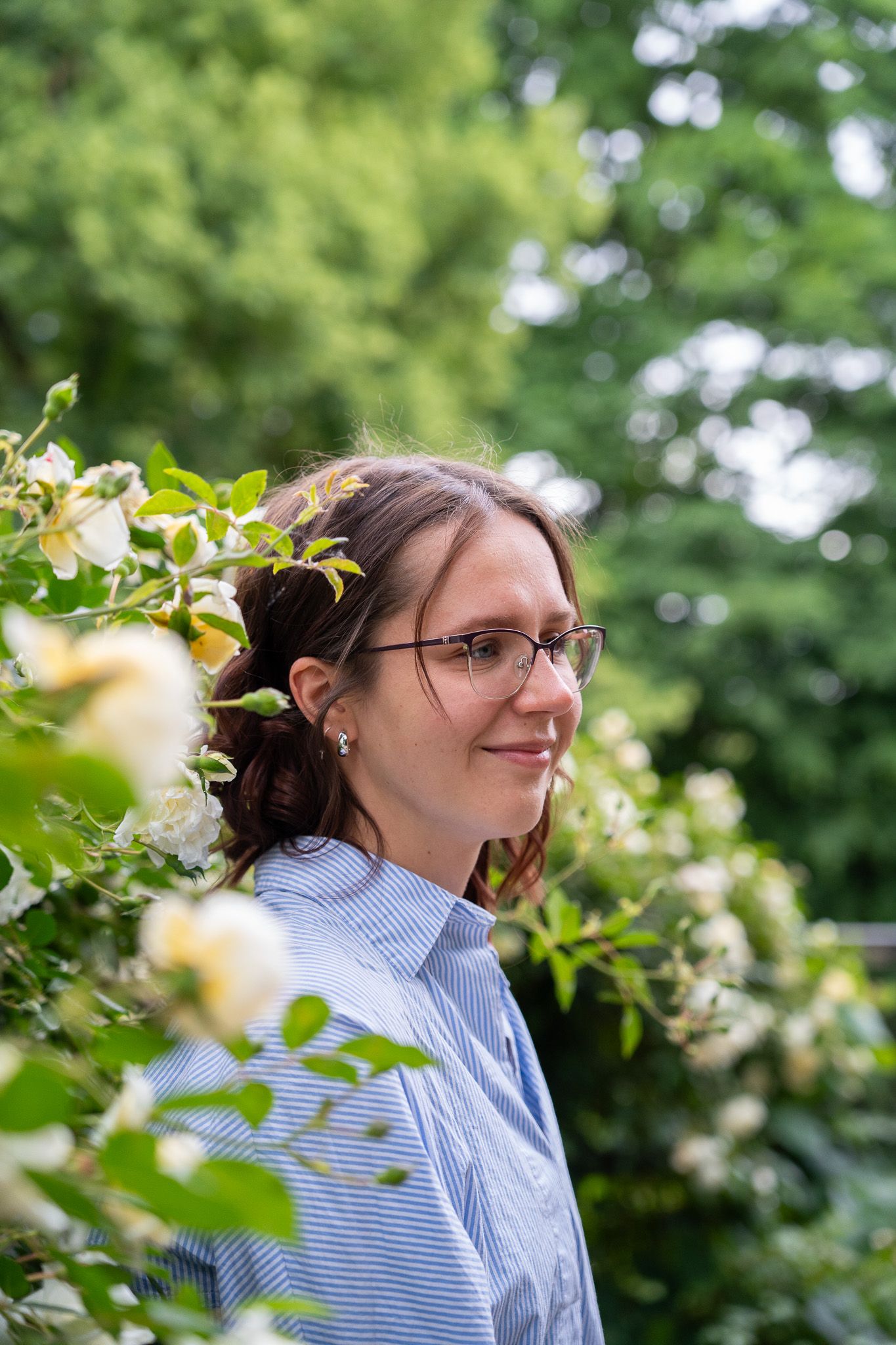 Young woman with glasses smiling softly surrounded by white roses and green foliage outdoors.