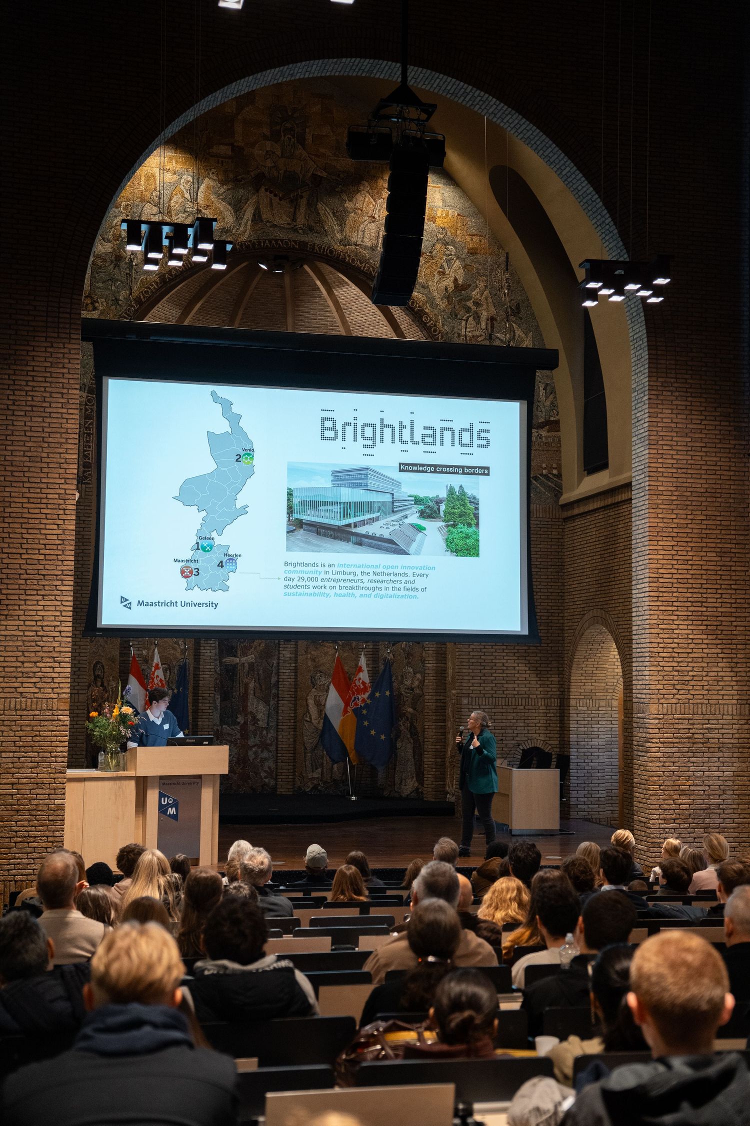 Speaker presents Brightlands campus information to a large audience in a grand Maastricht University hall.